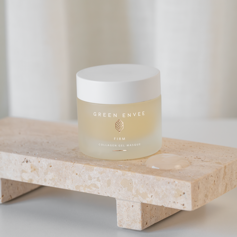 A frosted jar labeled 'Green Envee Firm Collagen Gel Masque' sitting on a beige stone slab with some gel beside it, in front of a light-colored curtain.