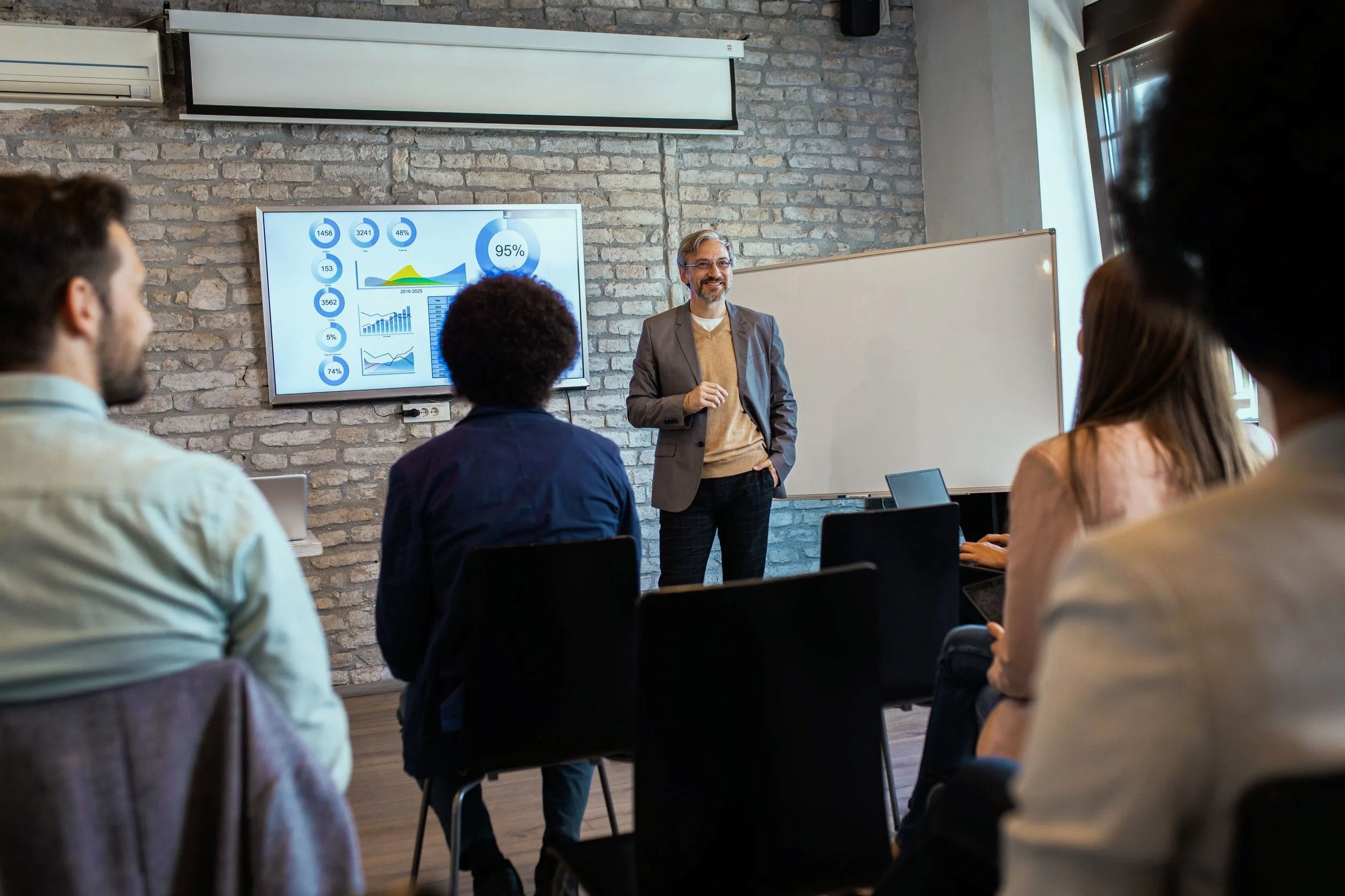A man in a blazer standing in front of a group of people giving a presentation in a conference room with a large screen displaying charts and data.