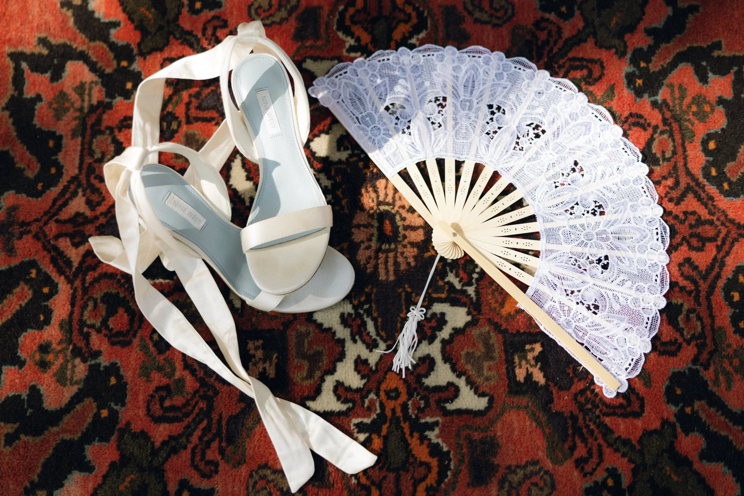 White lace fan and a pair of white high-heeled shoes with ribbon straps on a patterned carpet.