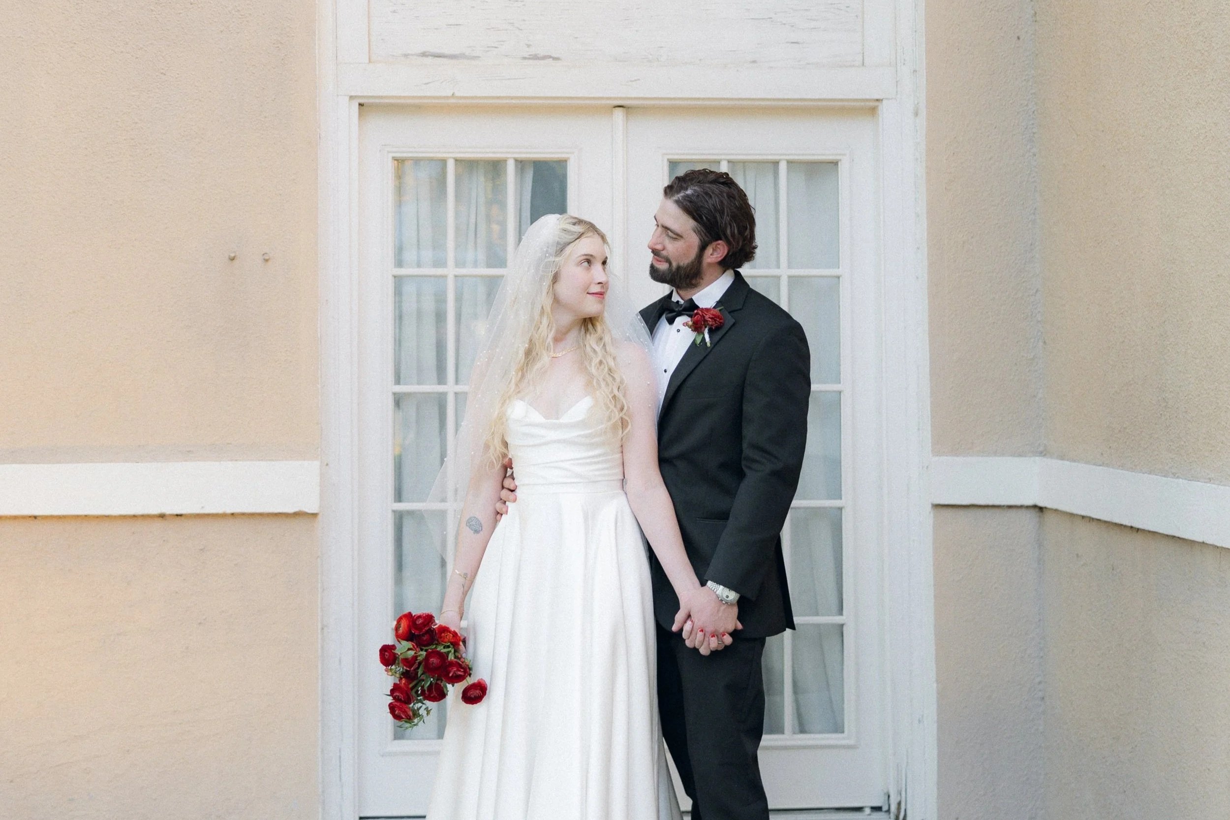 A bride and groom on their wedding day holding hands and looking at each other in front of a white door with glass panes.