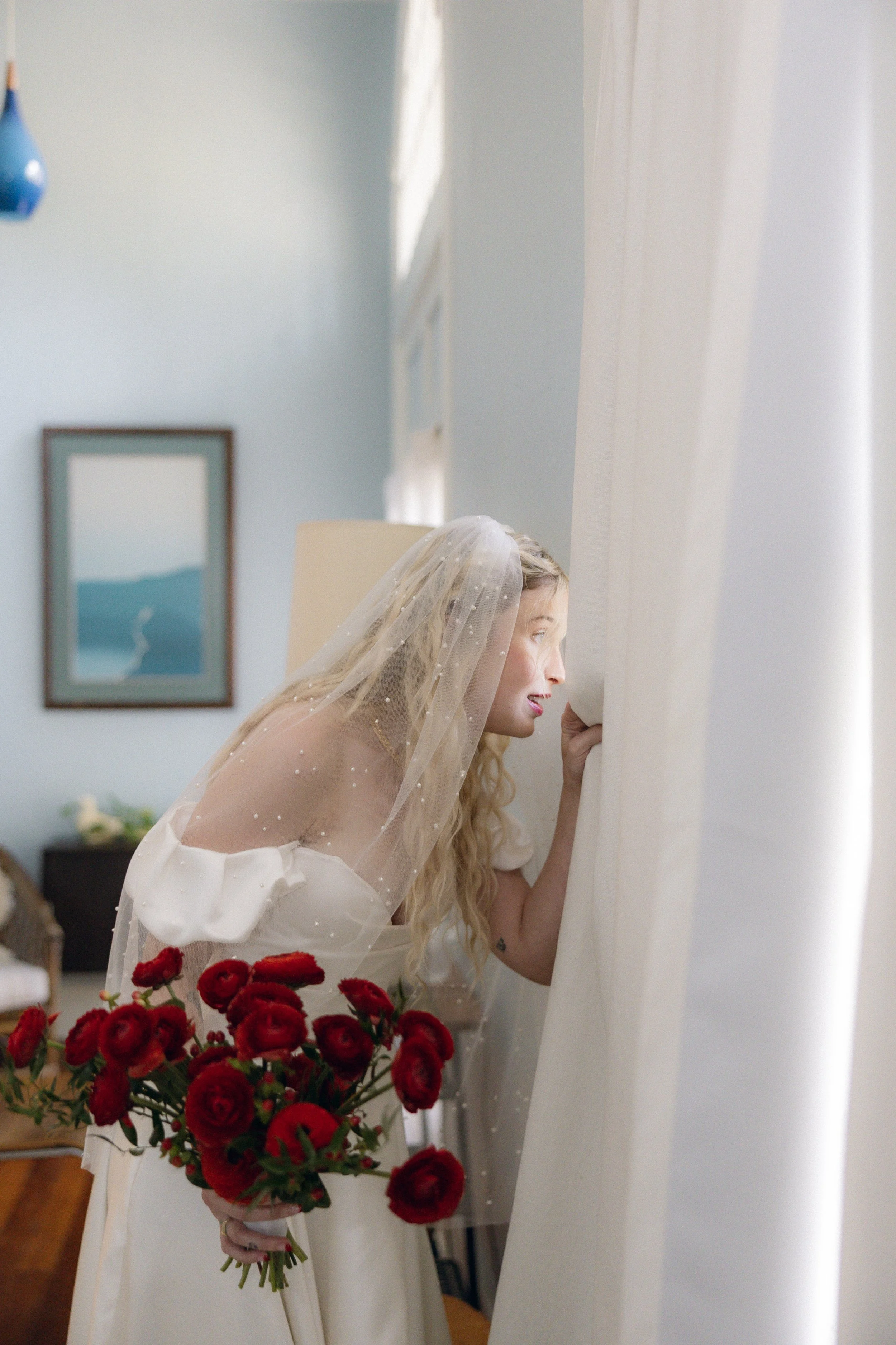 A woman dressed in a wedding gown and veil leaning over a window, holding a bouquet of red roses, looking out intently.