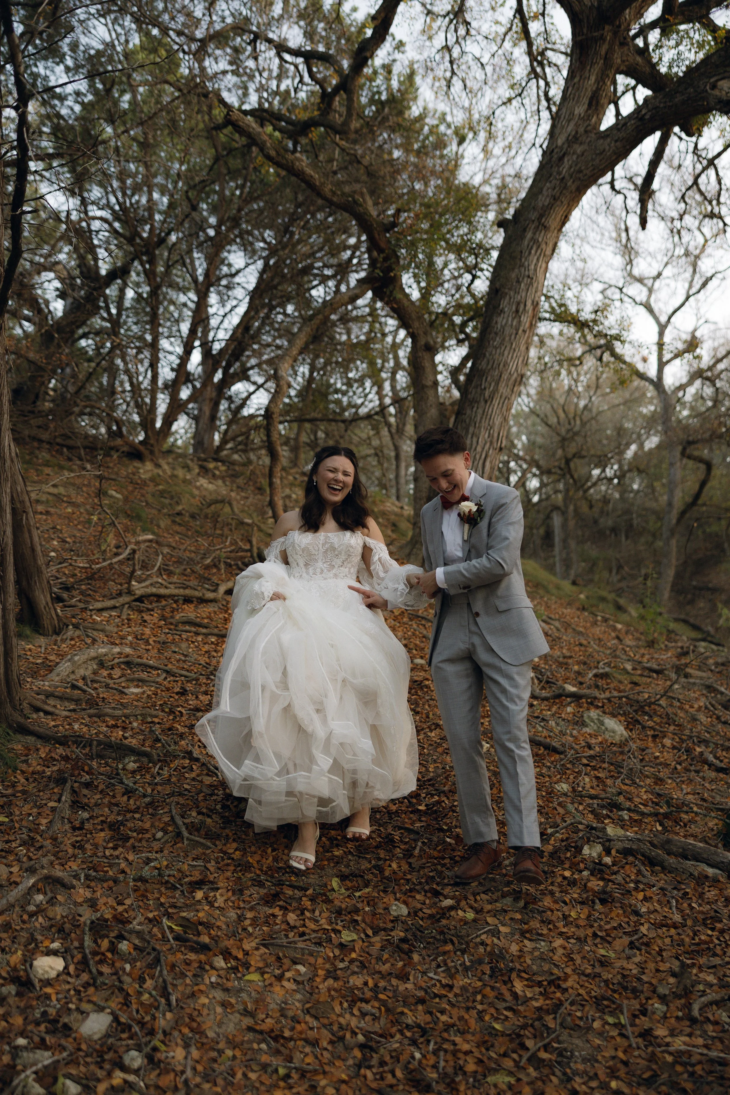 A newlywed couple walking through a wooded area with fallen leaves, sharing a joyful moment.