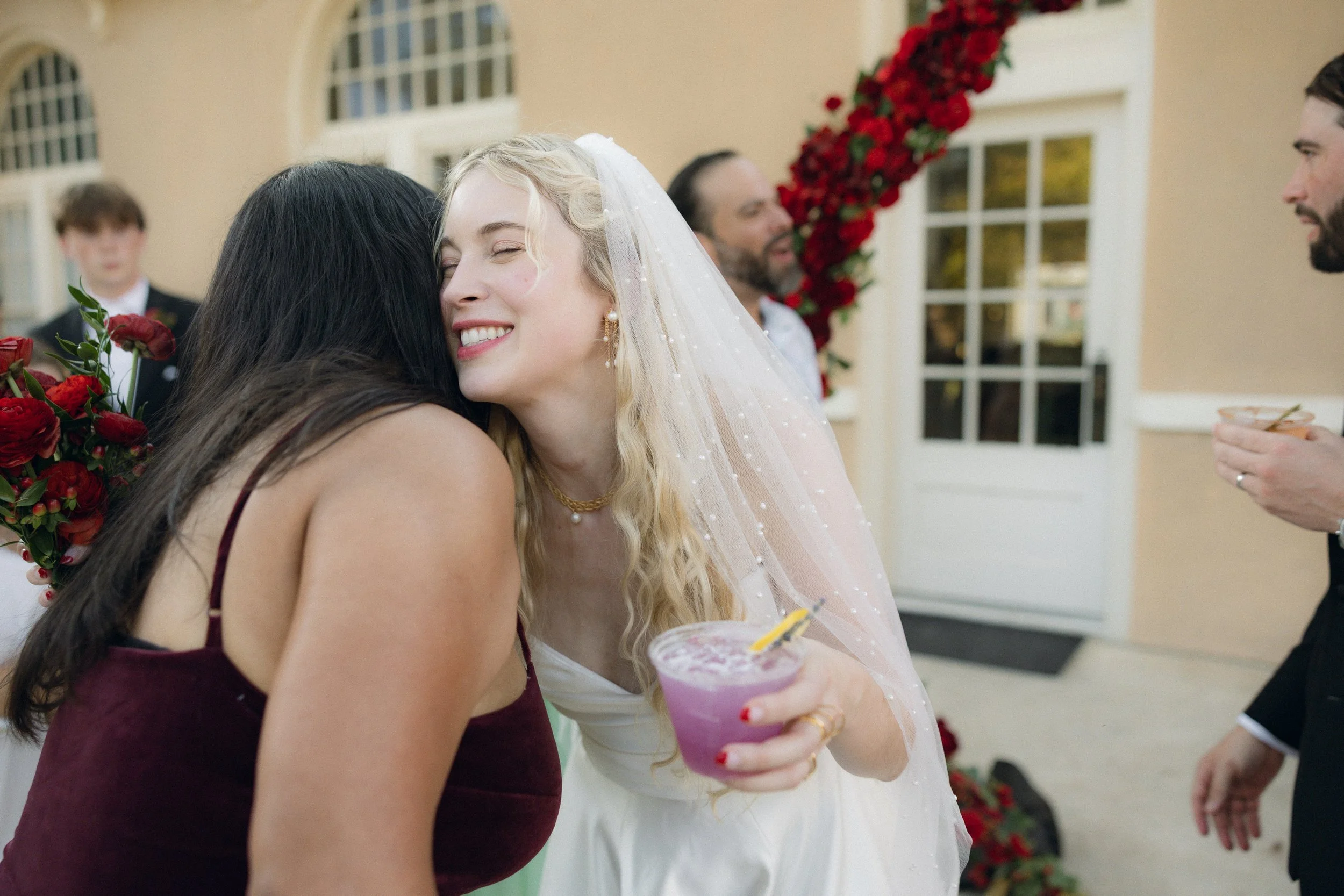A bride with long blonde hair and a white veil hugs a woman with long dark hair, who is holding a red flower bouquet, during a wedding celebration. Other guests are in the background, and there are red flowers decorating the area.