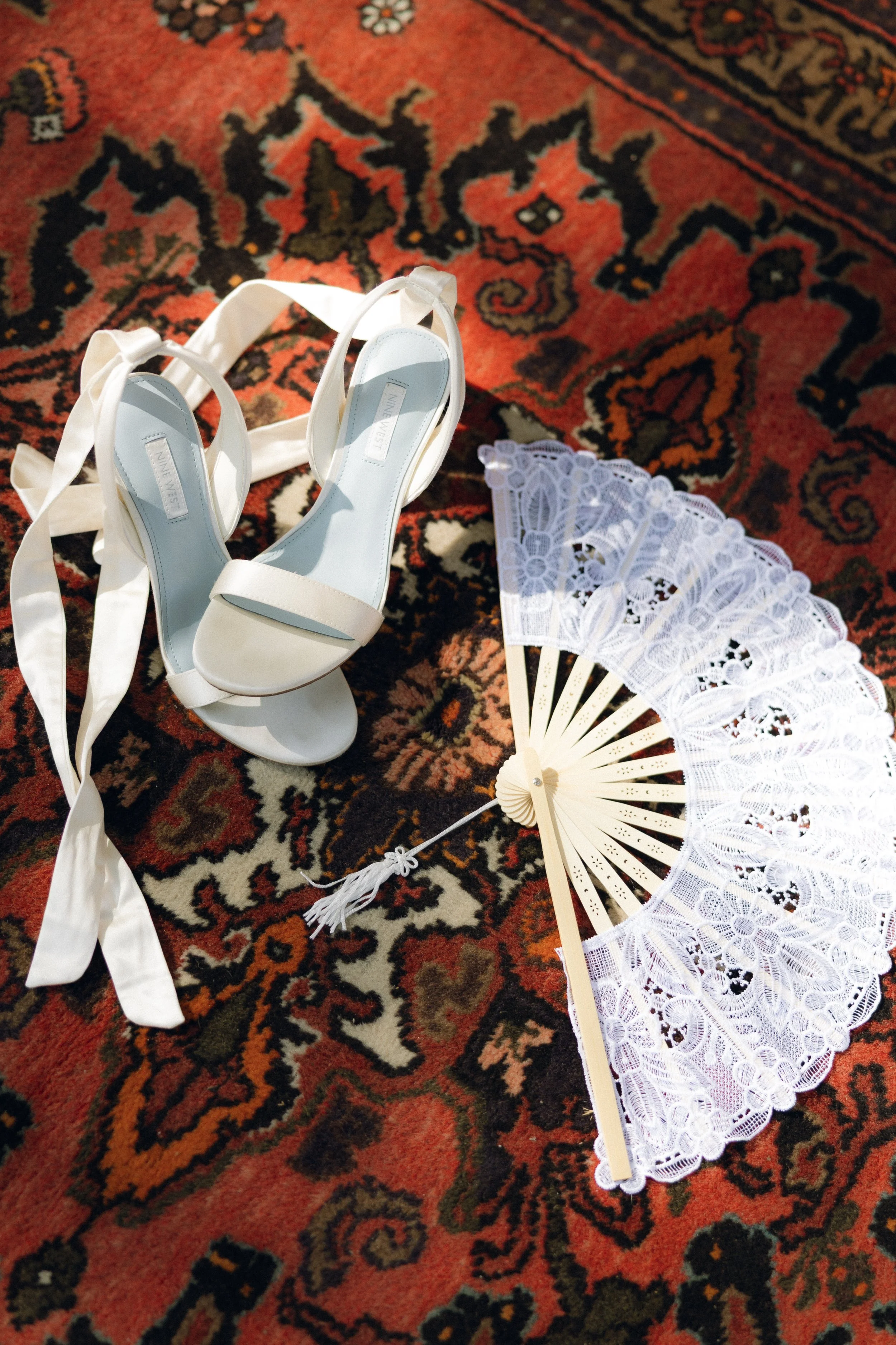white high-heeled shoes with ankle ribbons and a white lace folding fan on a decorative red and black rug.