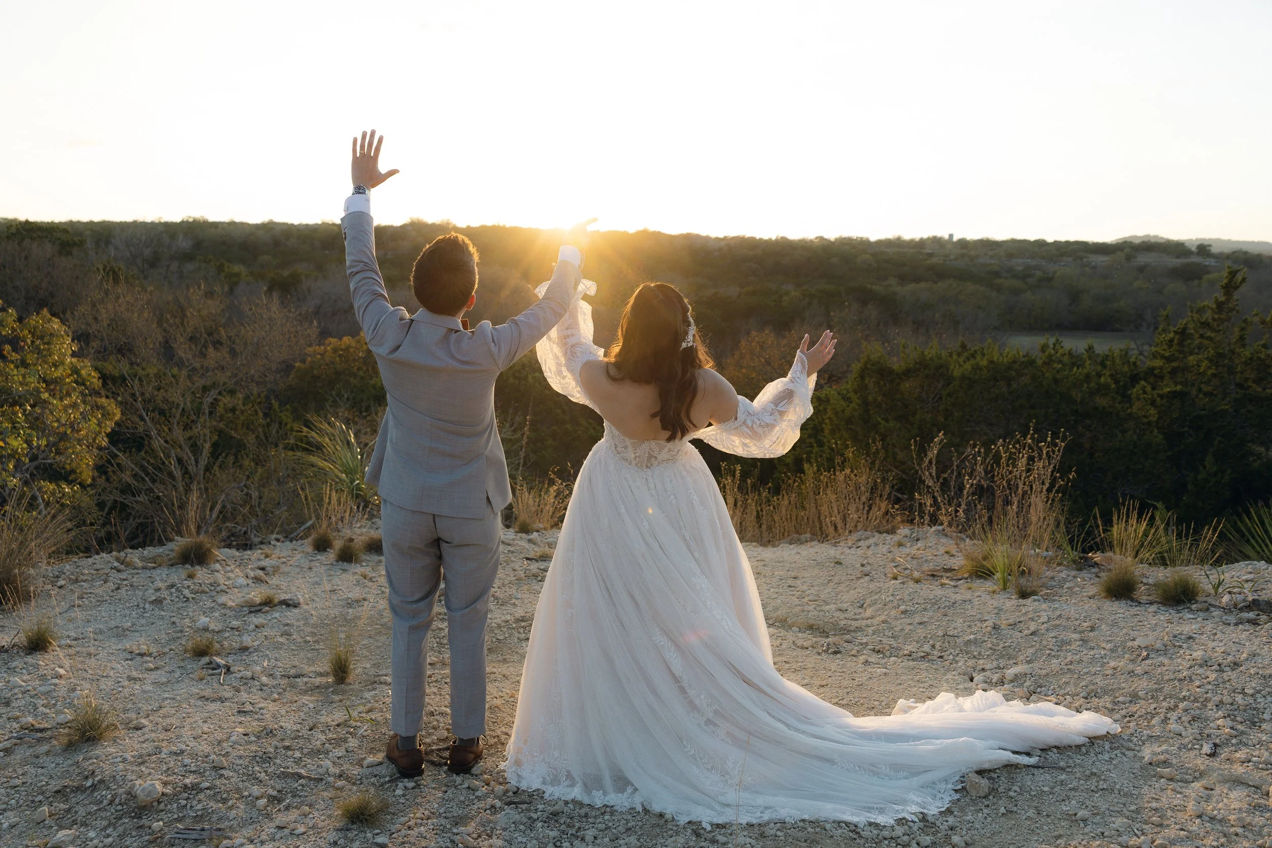 Two brides on their wedding day standing outdoors at sunset with arms raised, facing a scenic landscape of trees and hills.
