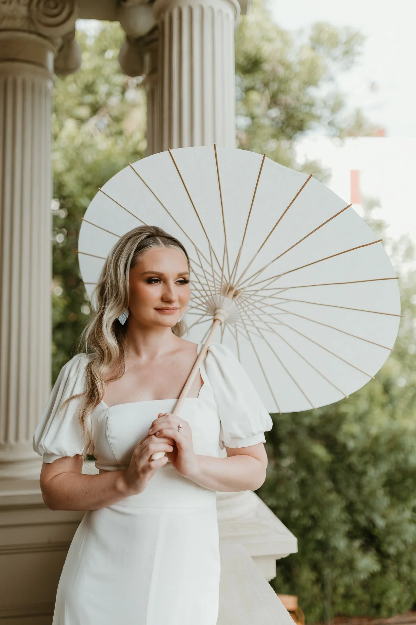 A bride at Hotel Ella in Austin, Texas holding a white parasol with a background of trees and classical columns.
