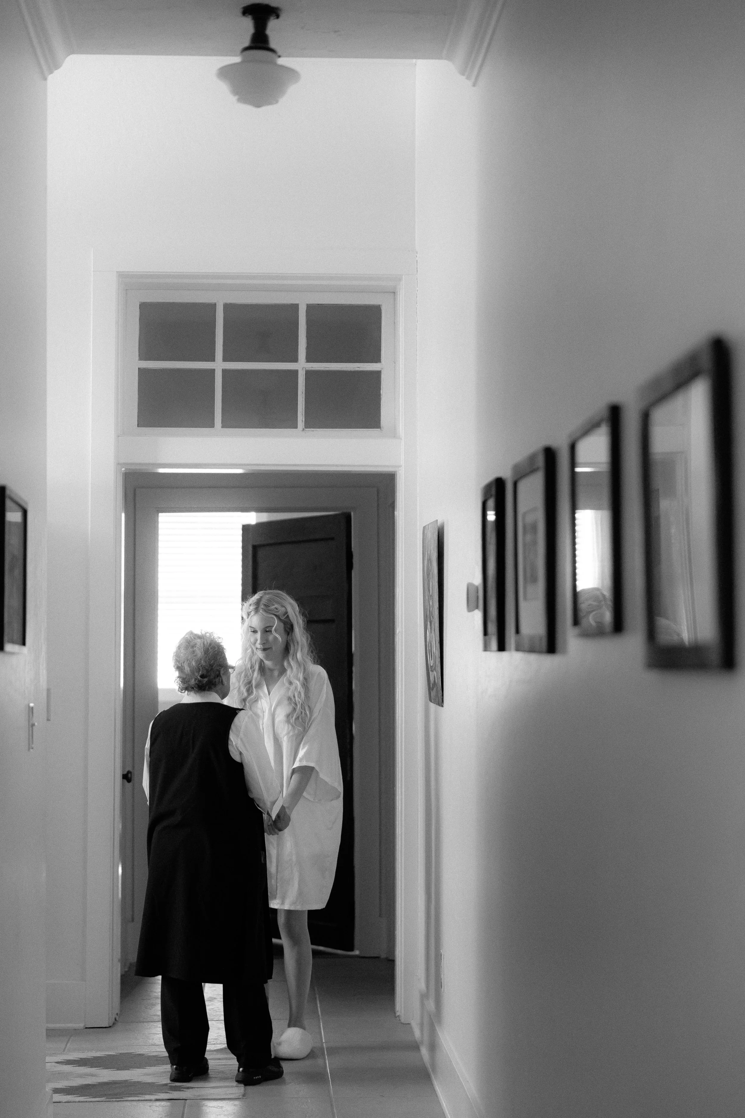 A bride in a white robe standing in a hallway, holding hands with an elderly woman dressed in black, with warm interaction and smiling faces.