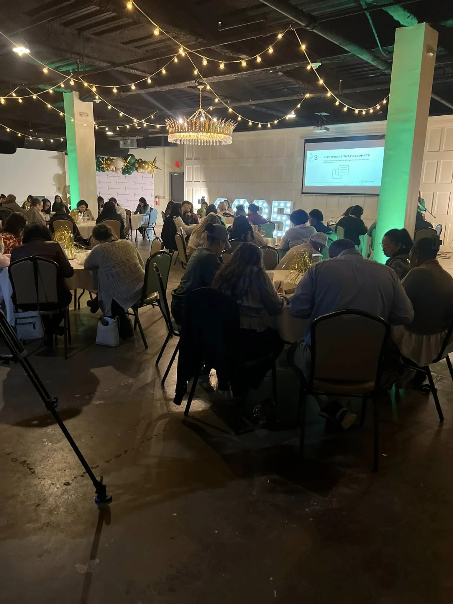 A conference or event space with people seated at round tables, a large screen displaying a presentation, decorative string lights overhead, and balloons and large illuminated numbers on the stage.