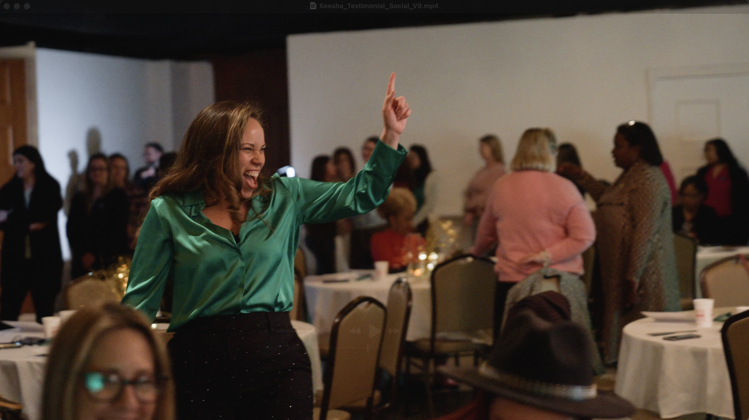 A woman with brown hair wearing a shiny green blouse and black pants is smiling and dancing at a social event, with several other people in the background near decorated tables.