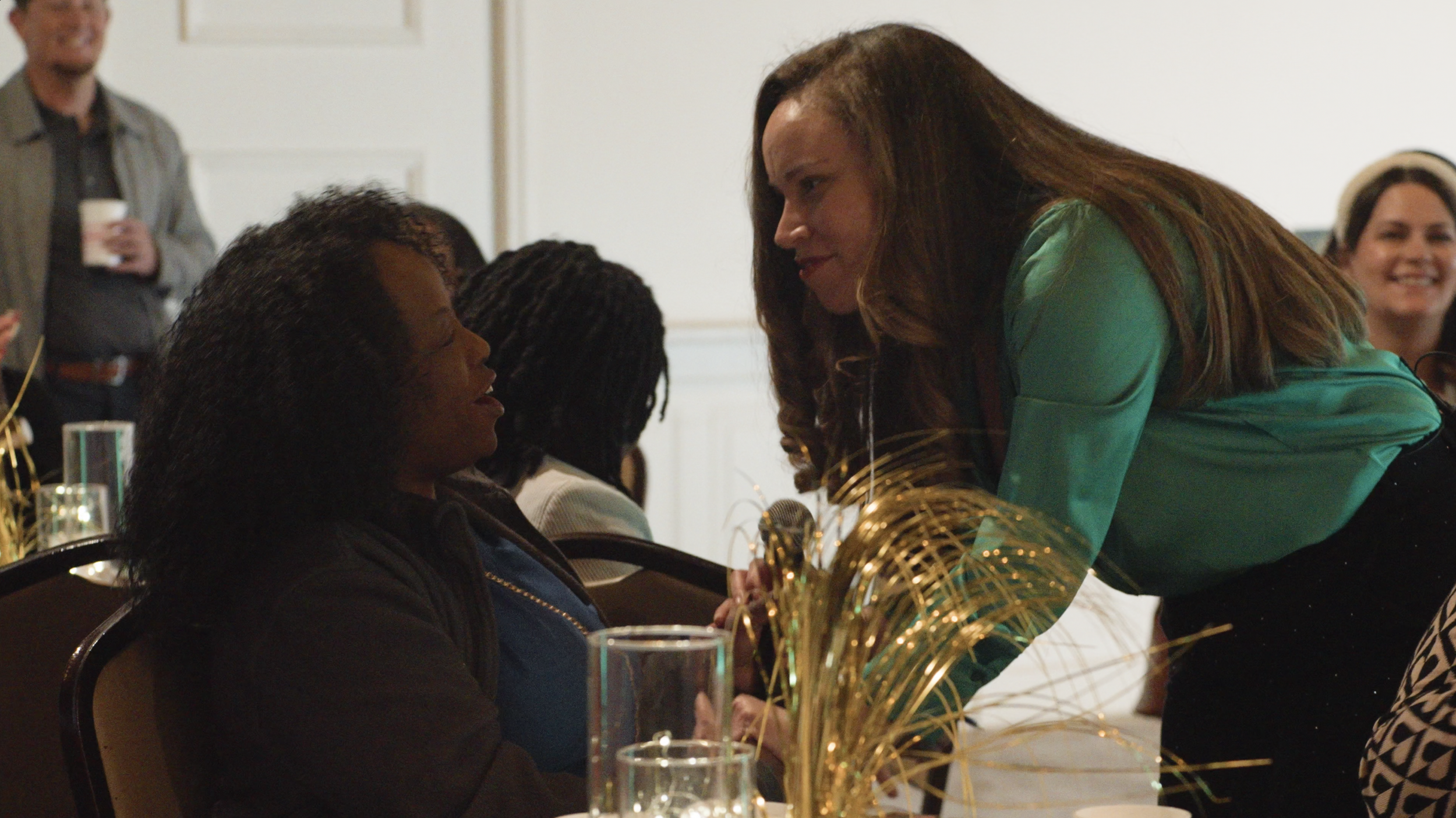 A woman with long brown hair wearing a green silk blouse leans over and speaks to a seated woman with curly black hair, during a social gathering or event.