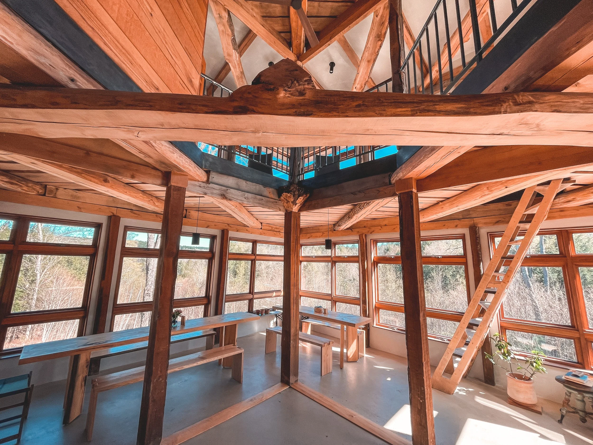 Interior of a bright, wooden two-story room with large windows, exposed beams, and a mezzanine level accessed by a ladder. Several wooden tables and benches are arranged inside, with sunlight streaming through the windows, revealing a view of trees outside.