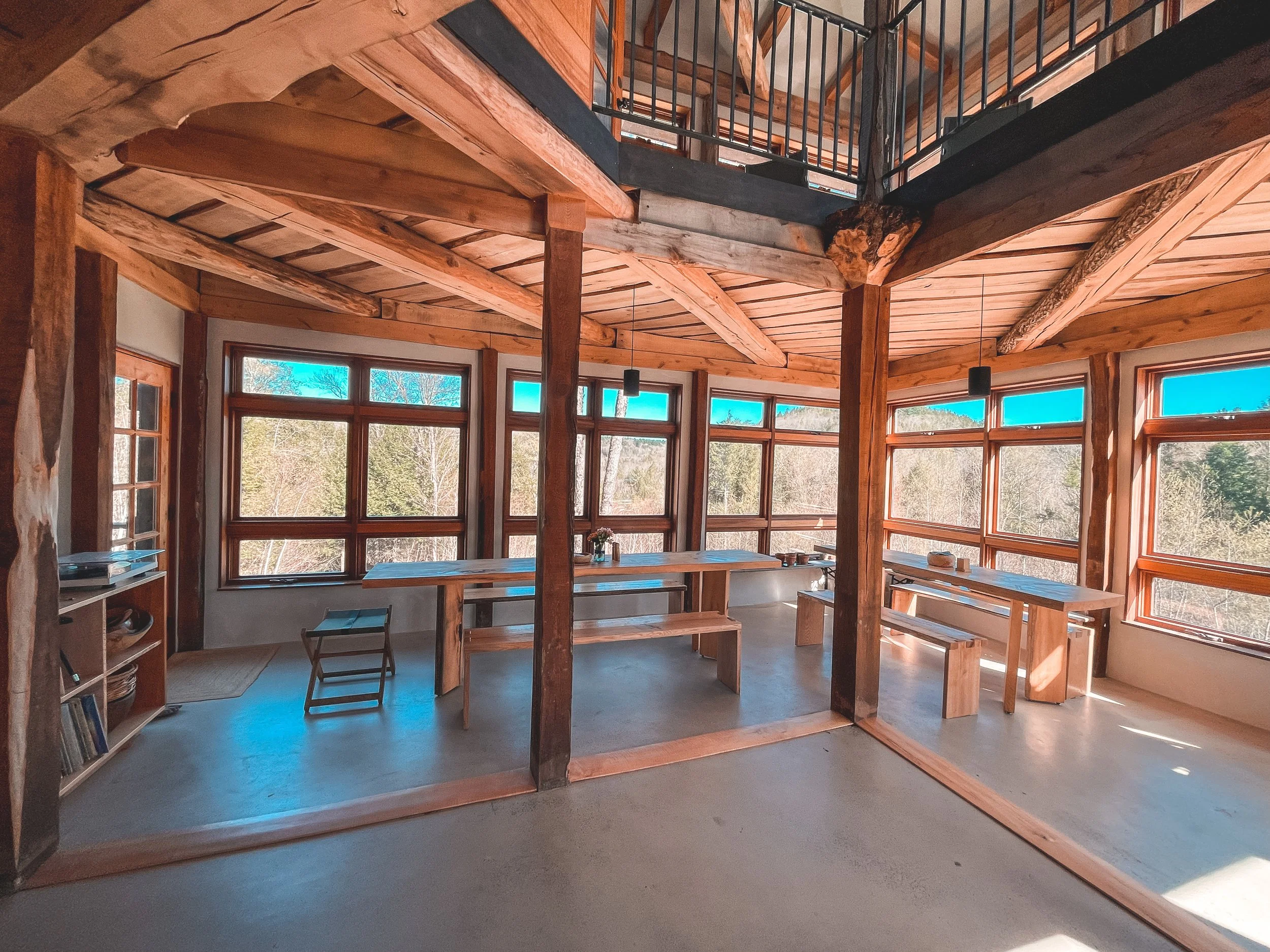 Sunlit room with large windows overlooking a wooded landscape, wooden beams and ceiling, minimalist furniture including tables, benches, and a small shelf with books.