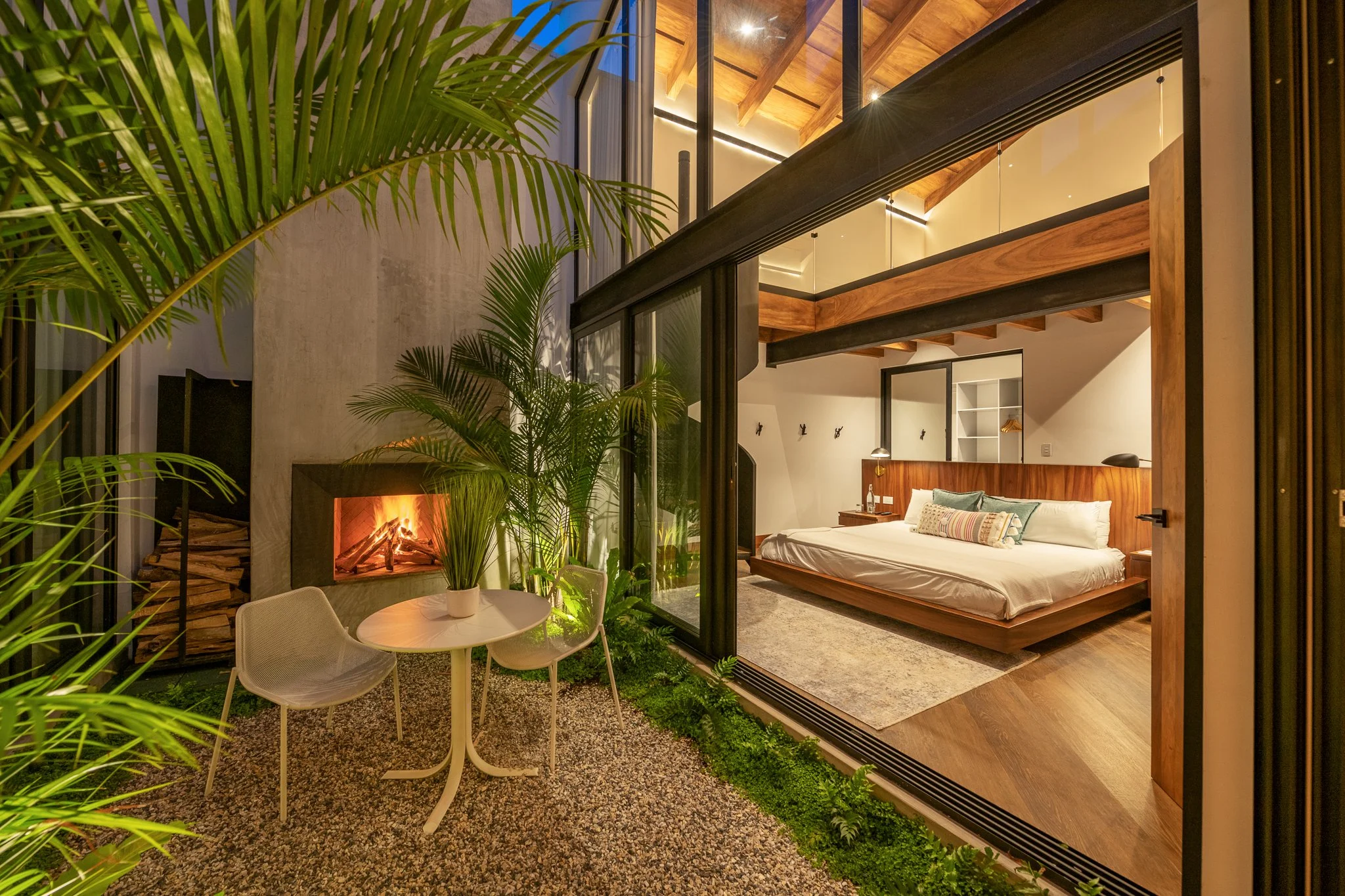 Interior de una habitación moderna con cama de madera, un área de estar con sillas y mesa, plantas verdes y una chimenea, con ventanas grandes que permiten vista al exterior Diseñado por Arquitectos en Antigua Guatemala Atelier