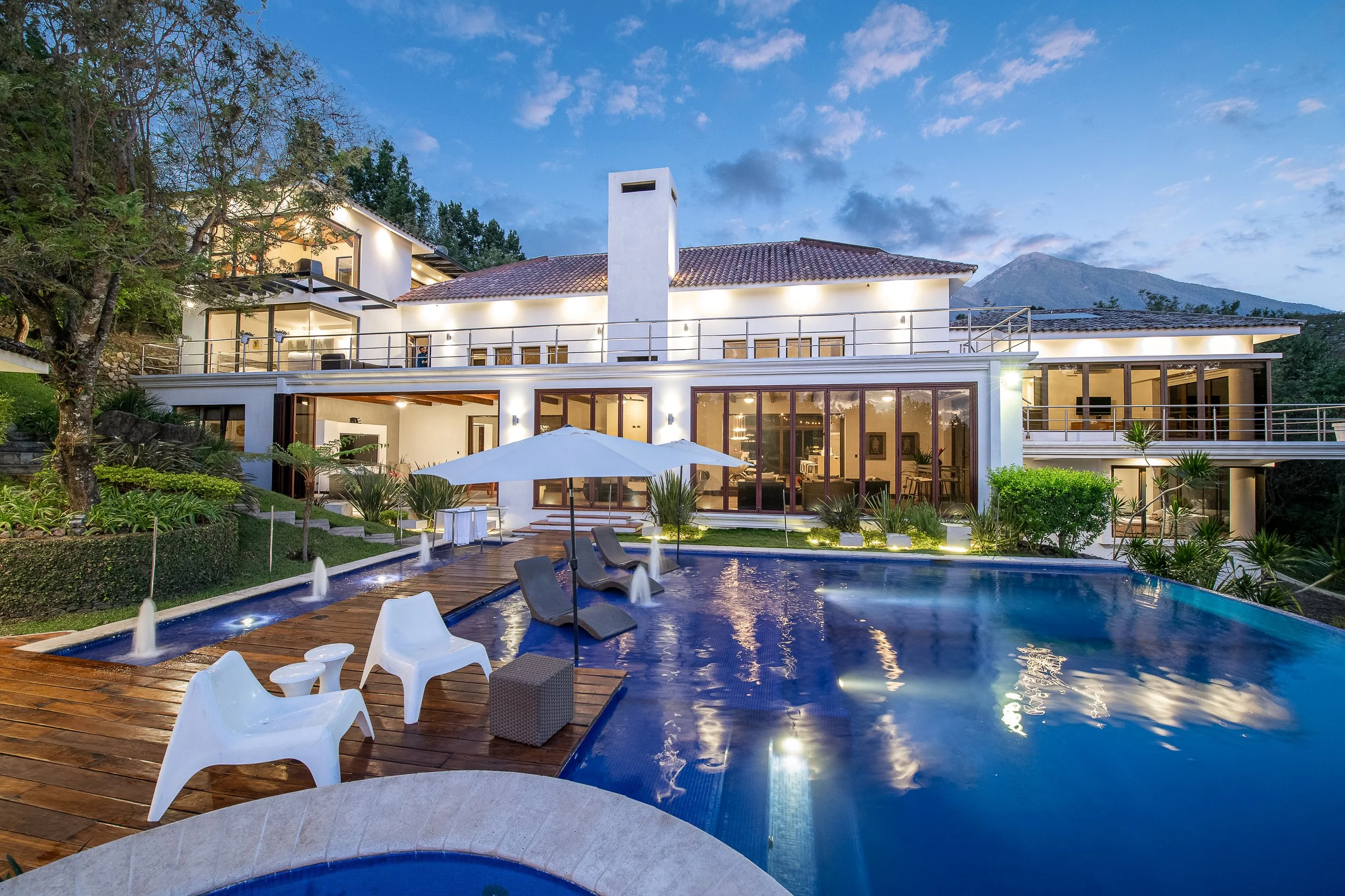Casa moderna con piscina y jardines en la noche, iluminación exterior, en un ambiente tranquilo y elegante con montañas al fondo.