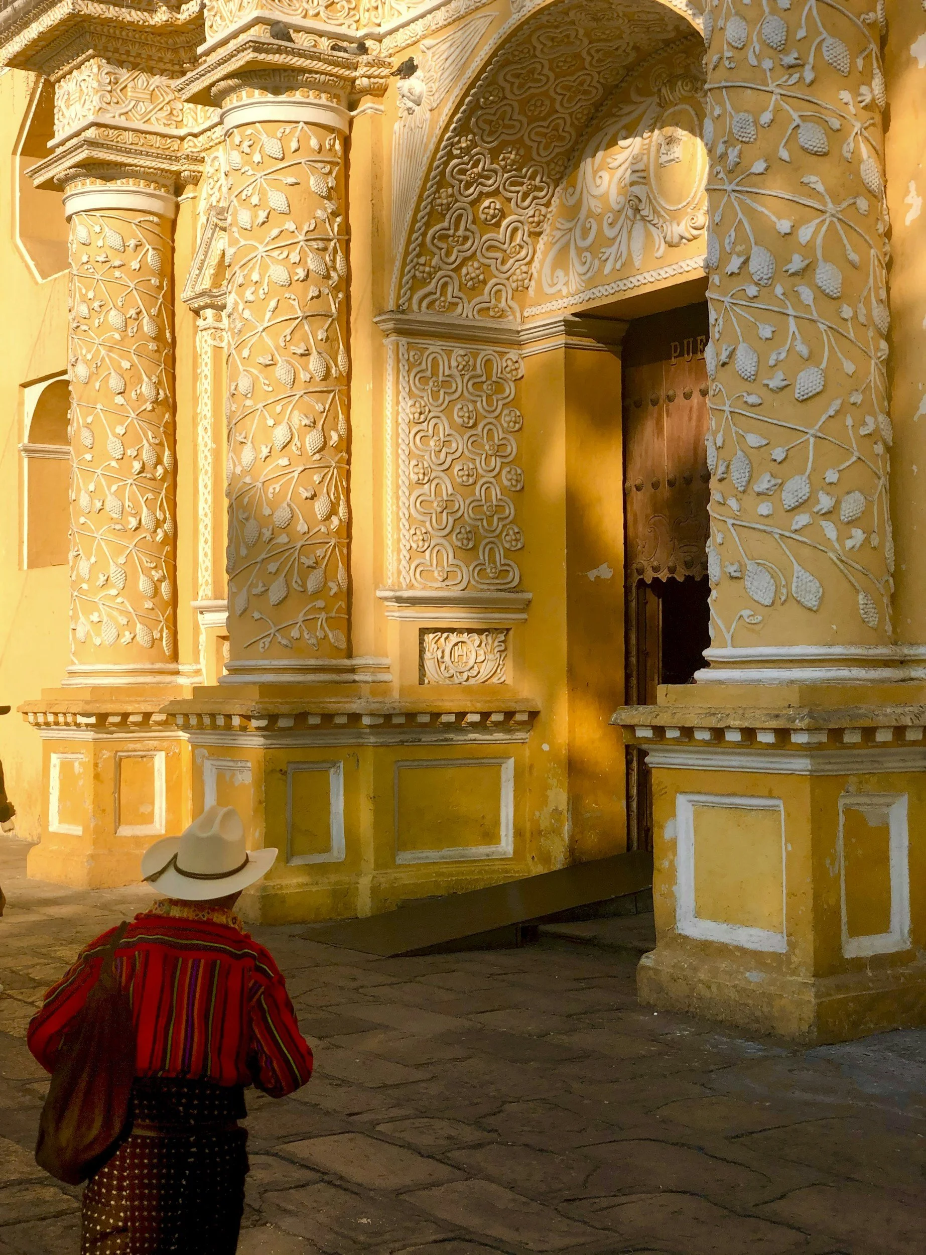Una persona con sombrero blanco y camisa roja con rayas verticales, caminando por una calle de piedra frente a un edificio colonial amarillo con detalles arquitectónicos decorativos en las columnas y arcos.