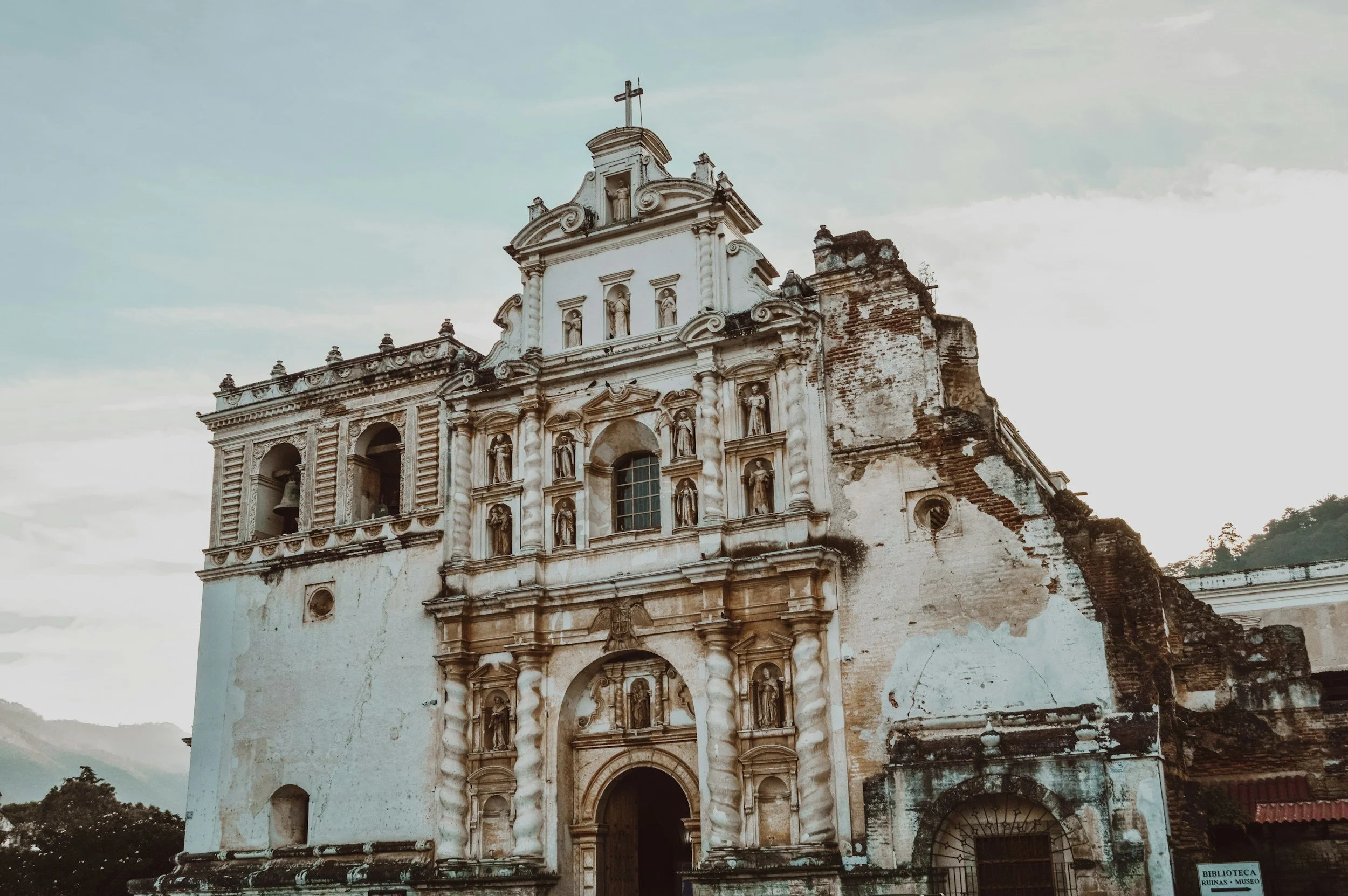 Fachada de una iglesia colonial en ruinas, con detalles ornamentales y una cruz en la cima, al atardecer.