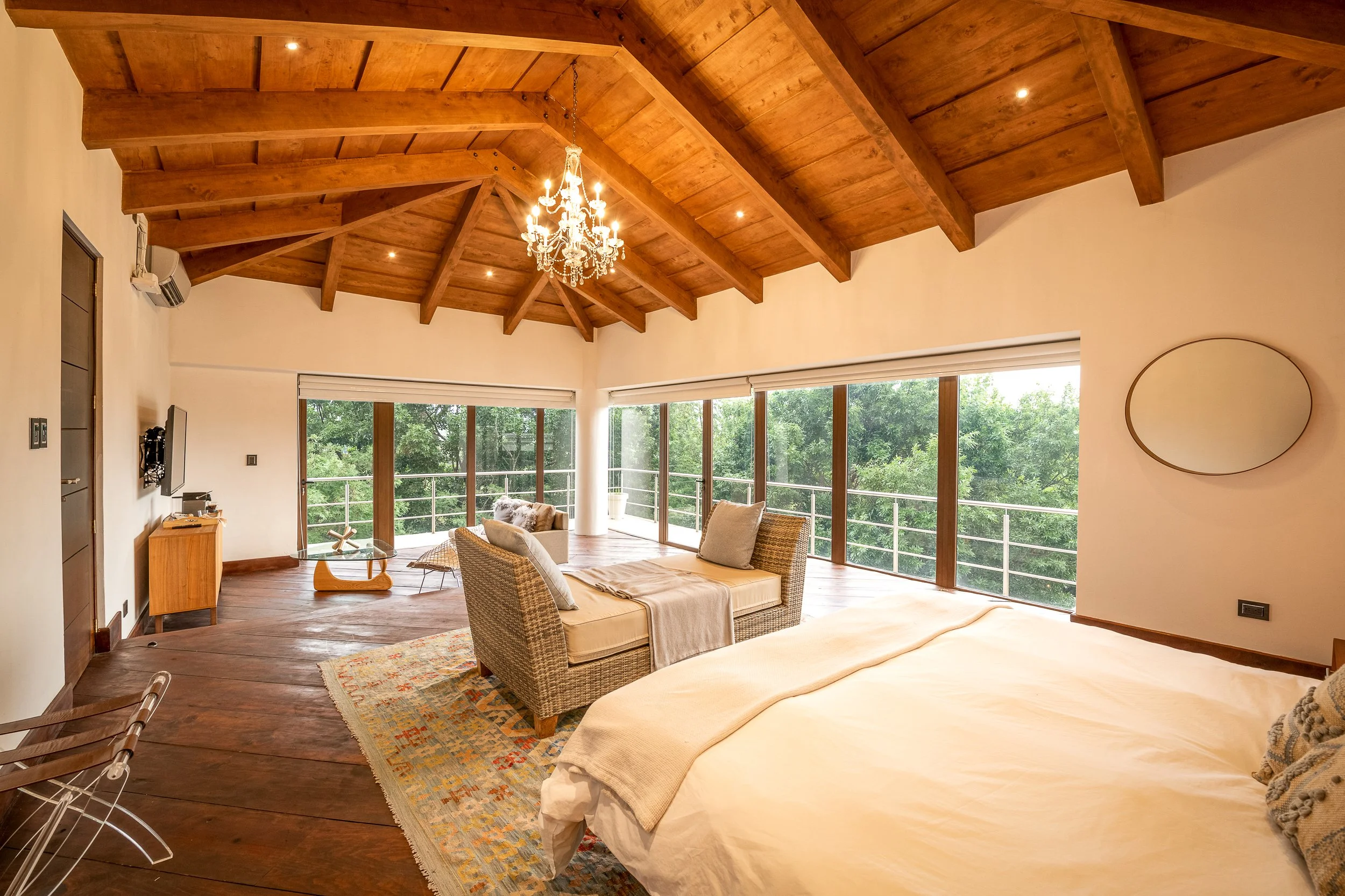 Habitat interior con techo de madera, cama, sillón, televisor y vistas a la naturaleza a través de grandes ventanas.