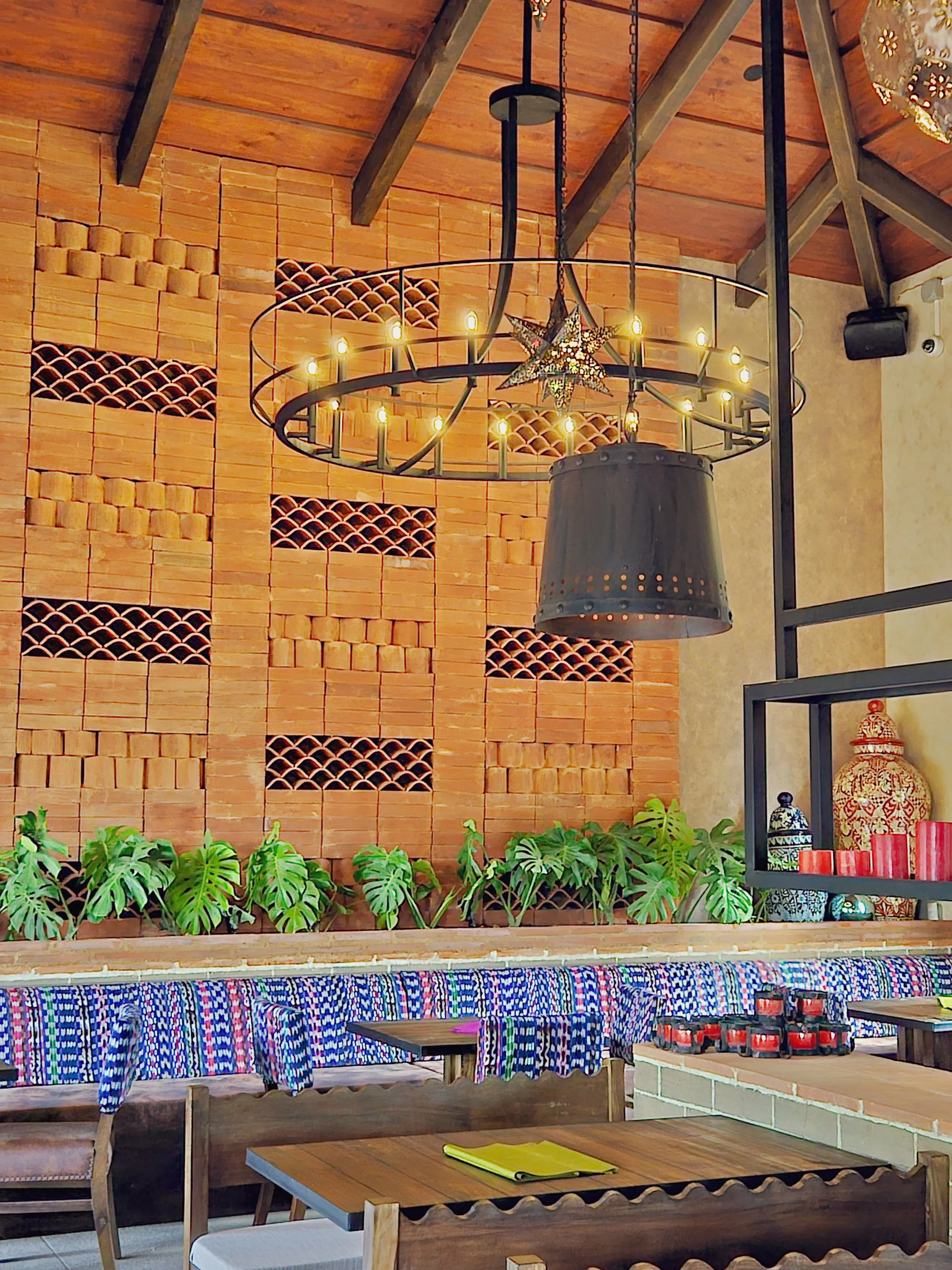 Interior de un restaurante con un mural de ladrillos en la pared, plantas verdes en una repisa, una lámpara de araña de hierro con velas y una decoración colorida con envases y velas en una estantería.