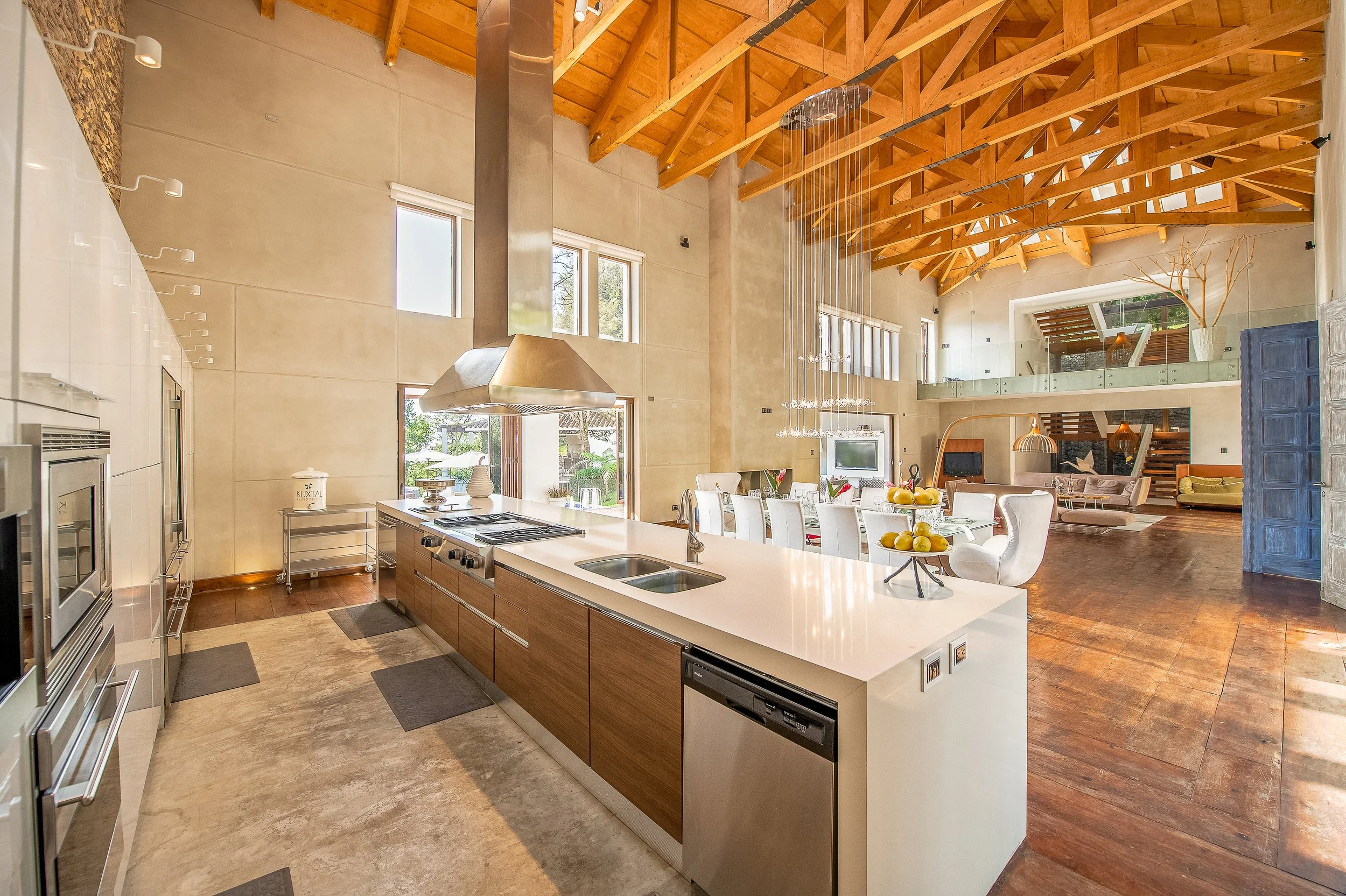 Interior de una sala de estar y cocina con techo de madera expuesta y ventanas altas, comedor con sillas blancas y sala con sillones, decoración moderna y luminosa.