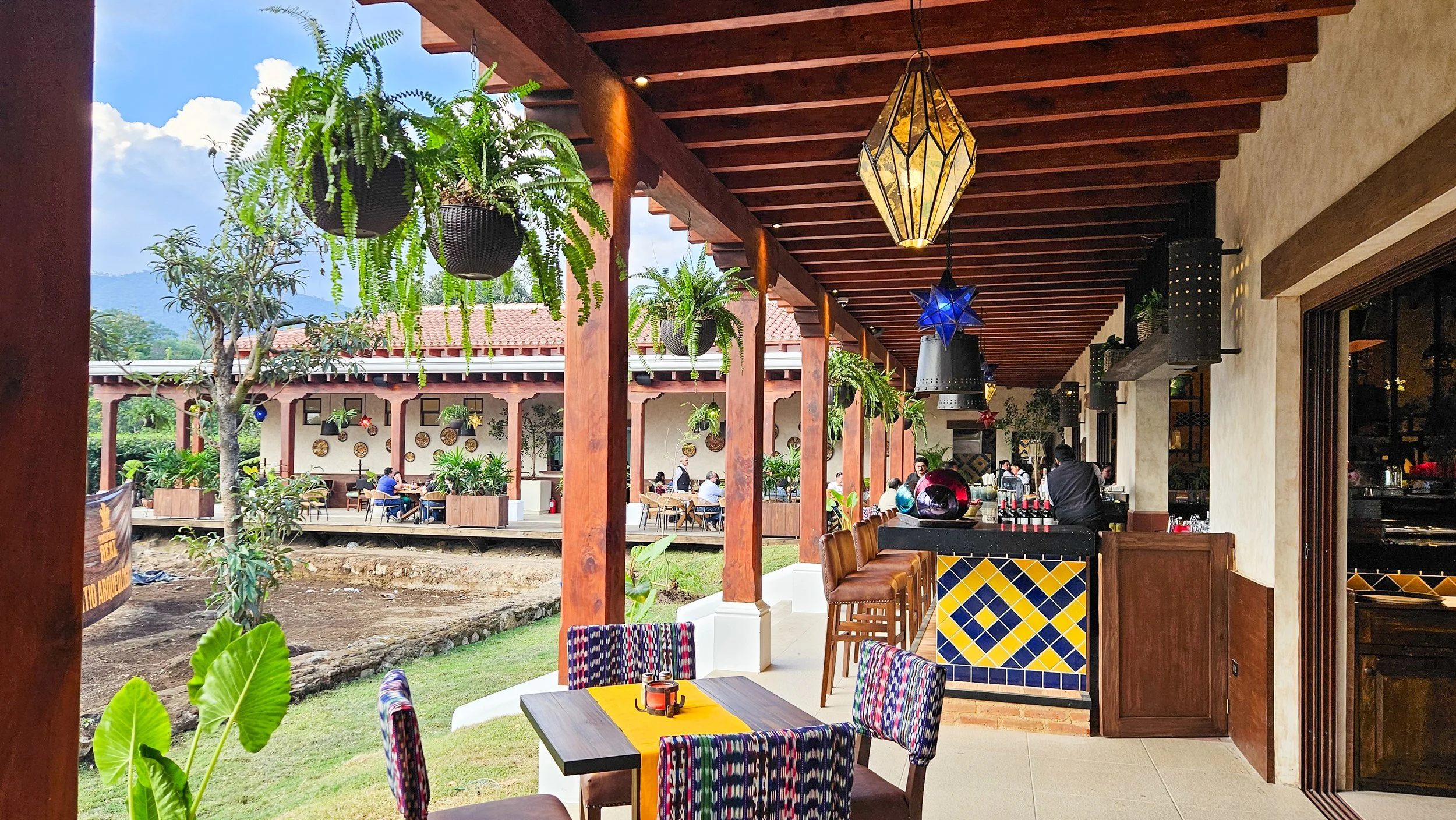 Patio exterior de restaurante con mesas y sillas, plantas en colgantes y paredes decoradas, vista hacia un área de comedor al aire libre con más mesas y plantas, cielo despejado con nubes.