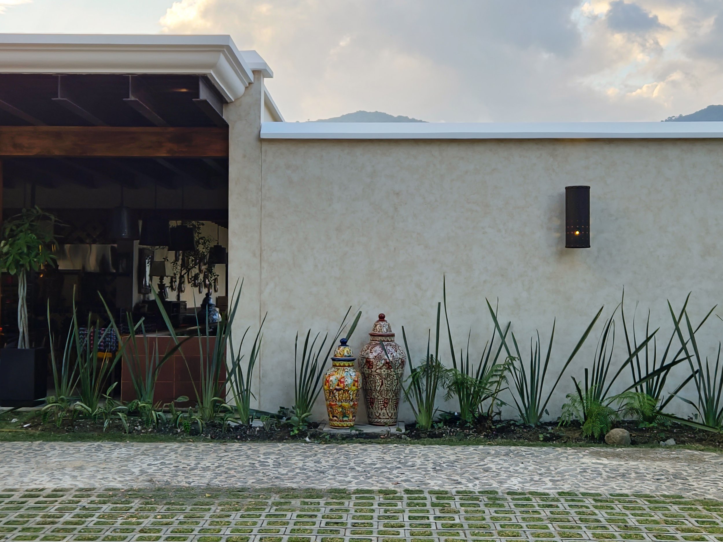 Vista exterior de un muro beige con decoraciones de cerámica y plantas verdes, en un entorno de paisaje natural y con cielo nublado.