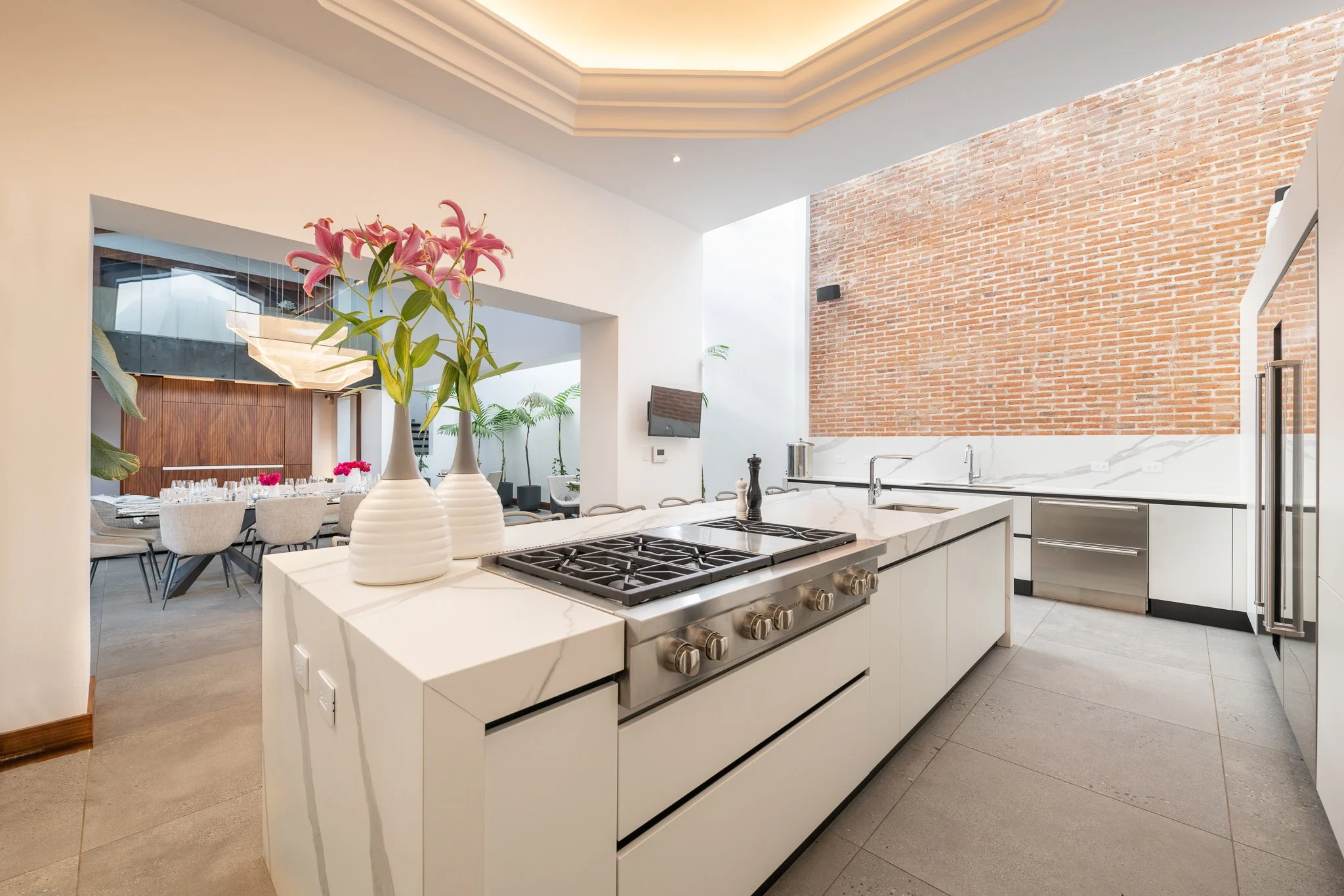 Cocina moderna con isla central blanca, estufa de gas, paredes de ladrillo expuesto y diseño abierto con comedor visible al fondo con sillas beige y flores rosas Diseñado por Arquitectos en Antigua Guatemala Atelier