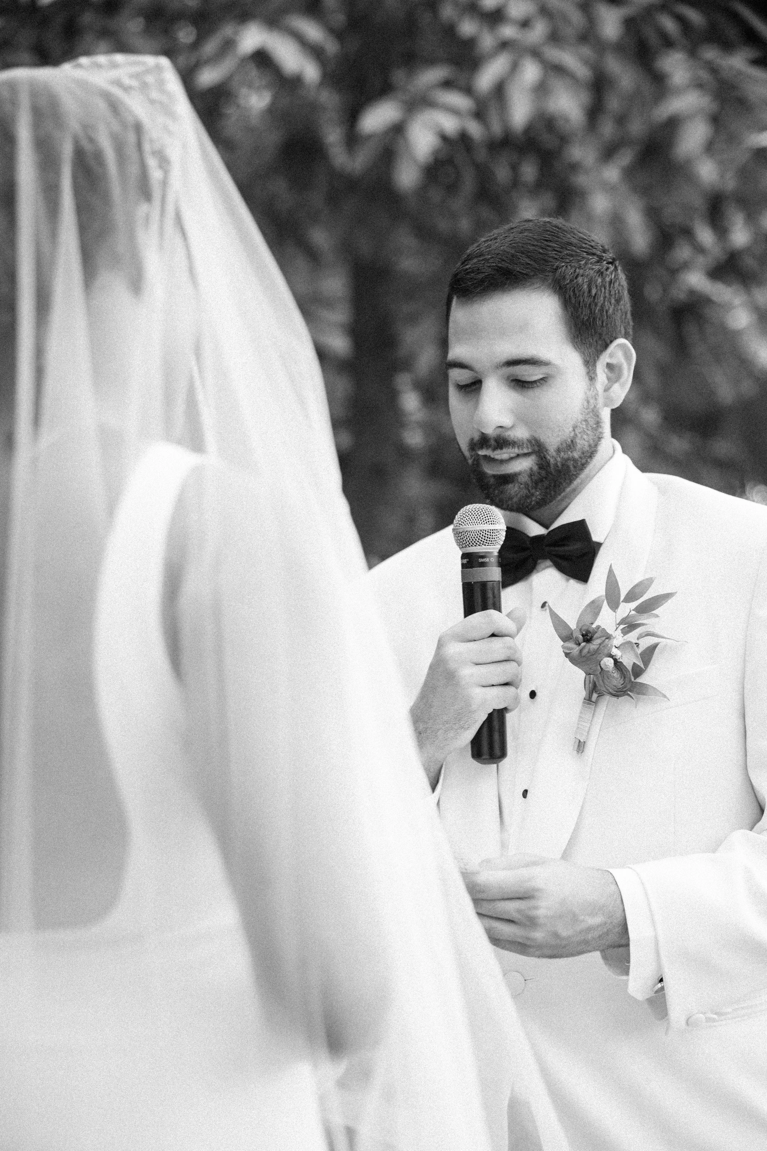 A man in a tuxedo holding a microphone, likely at a wedding ceremony, with a woman in a wedding dress in the foreground.
