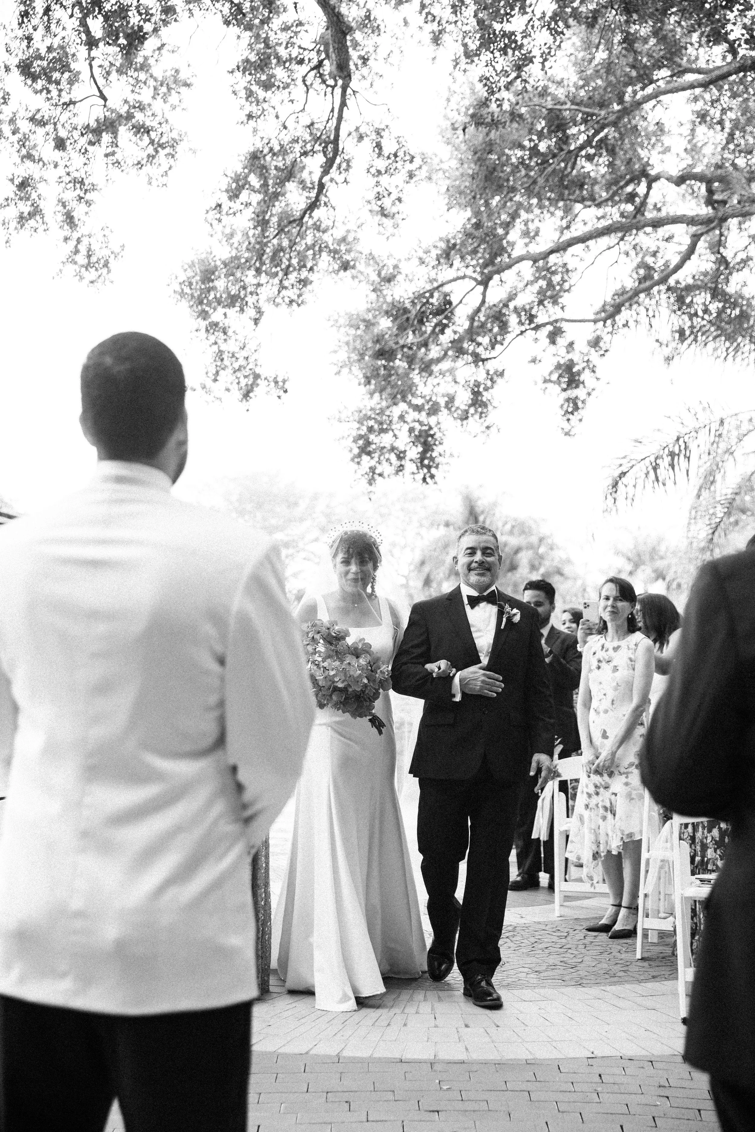 Black and white photograph of a wedding ceremony outdoors, with the bride and an older man, possibly her father, walking down the aisle. The bride is holding a bouquet, and guests are watching and smiling.