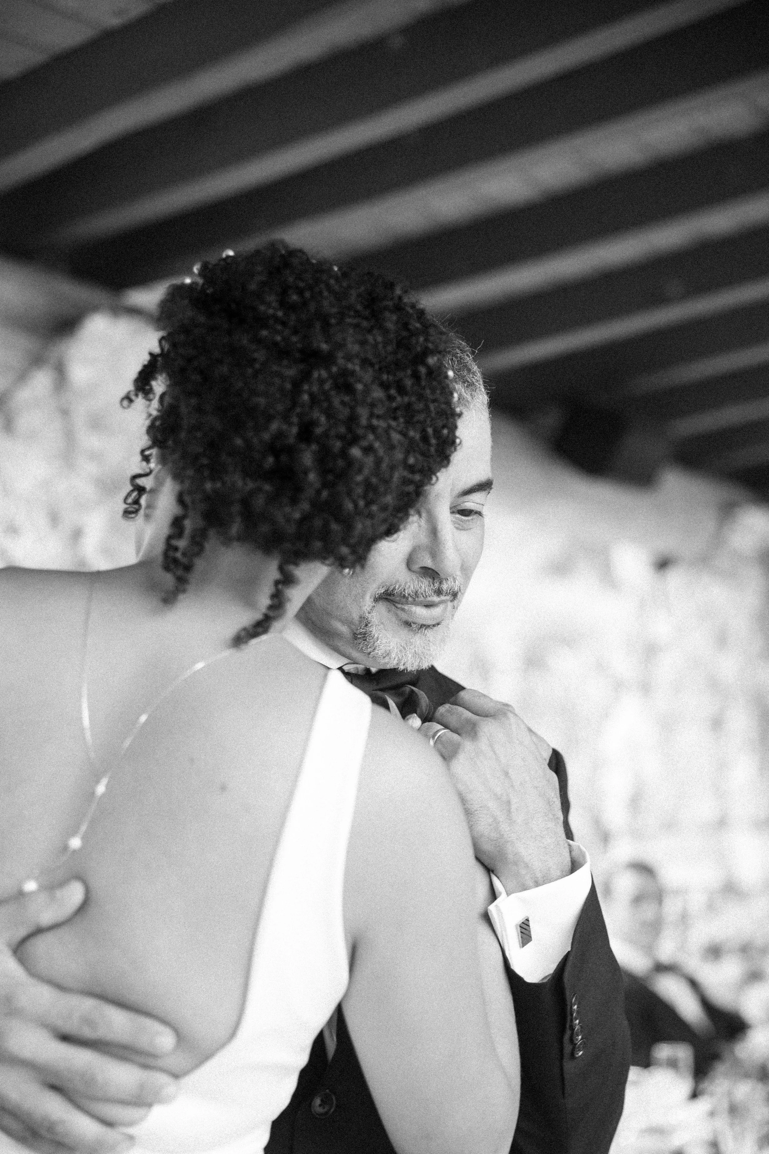 A black and white photo of a man and a woman embracing, with the man adjusting his tuxedo collar and the woman’s face partly visible. They appear to be at a formal event or celebration, likely dancing or sharing an intimate moment.