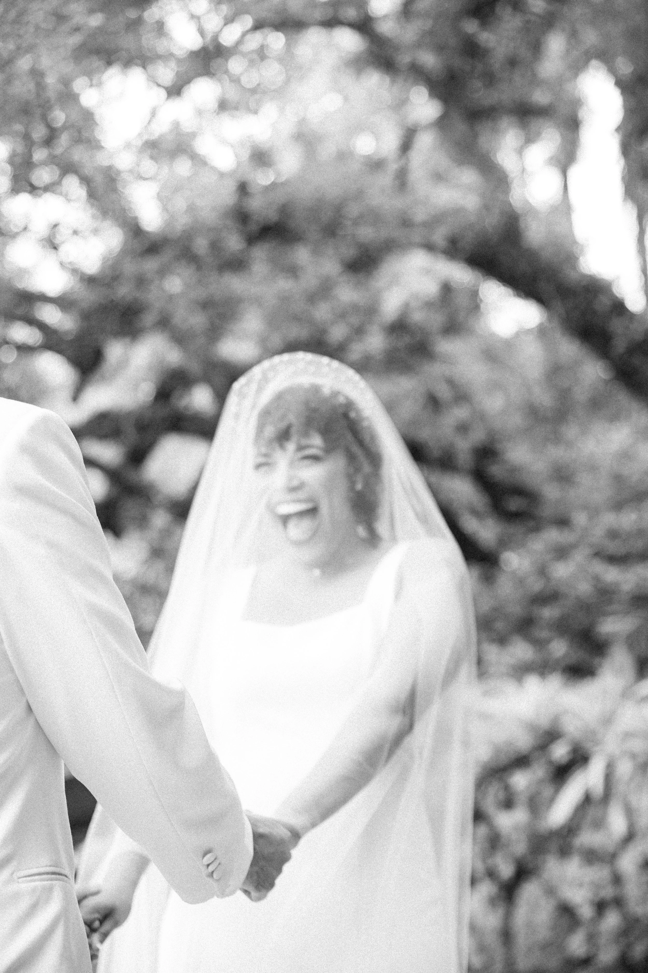A bride in a wedding dress with a veil smiling joyfully while holding hands with her groom during an outdoor wedding.
