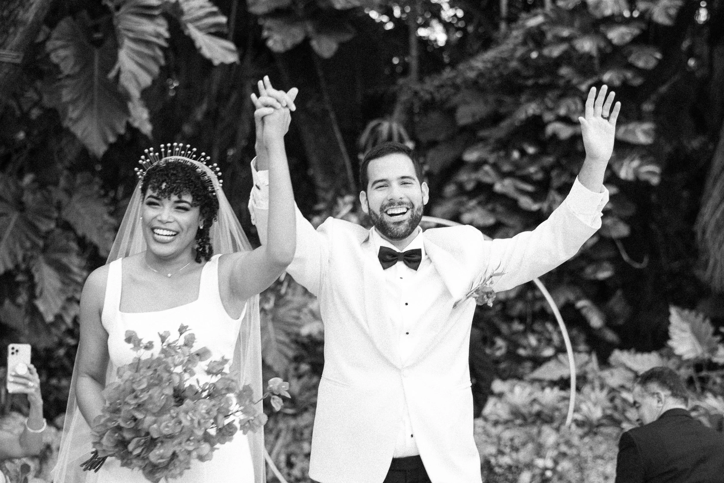 A bride and groom celebrating at their wedding, holding hands with arms raised, smiling happily. The bride is wearing a wedding dress with a veil and crown, and the groom is dressed in a tuxedo with a bowtie. They are outdoors with large leaves in th