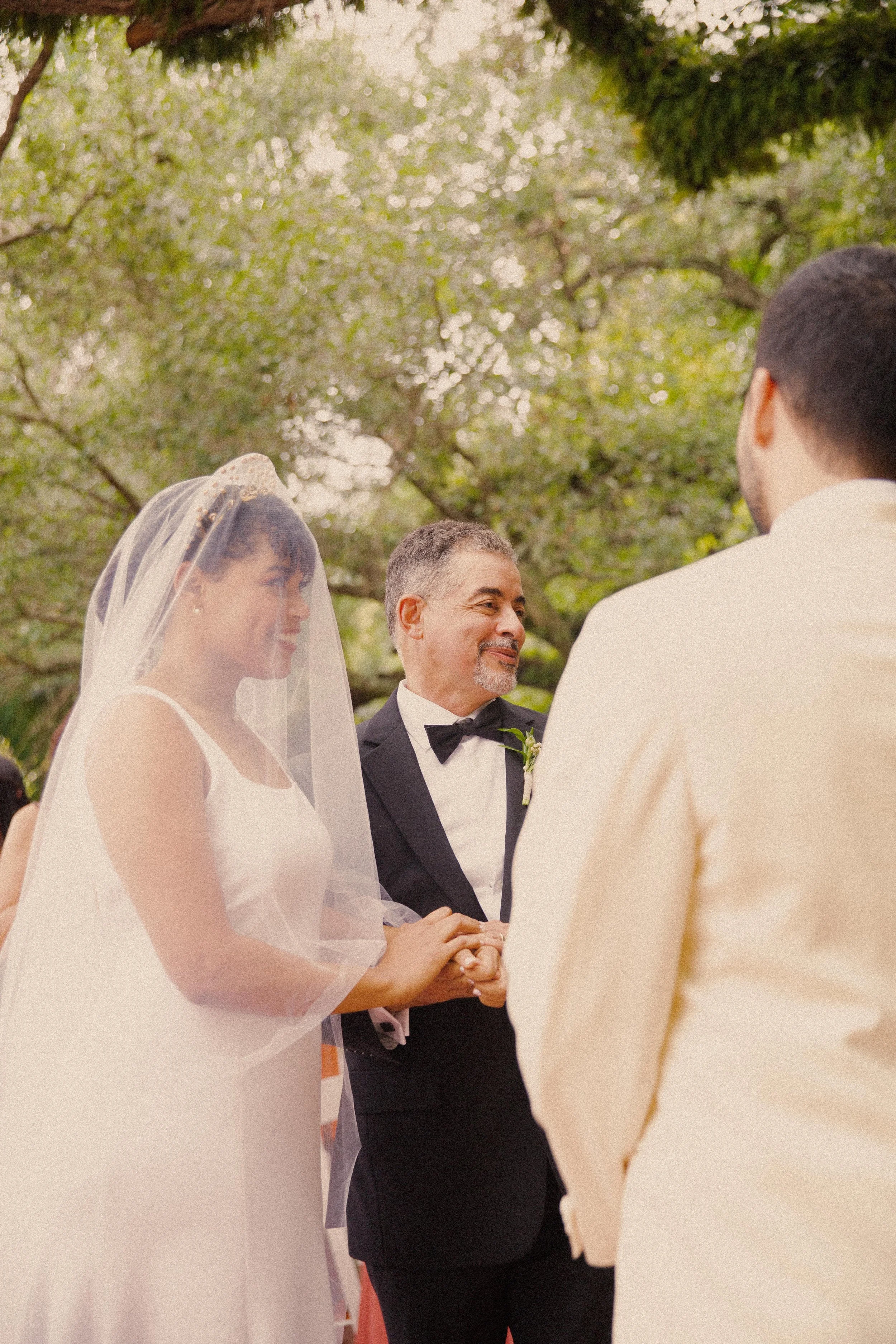 Bride and groom holding hands during a wedding ceremony outdoors, with a man in a tuxedo officiating, under trees with green leaves.