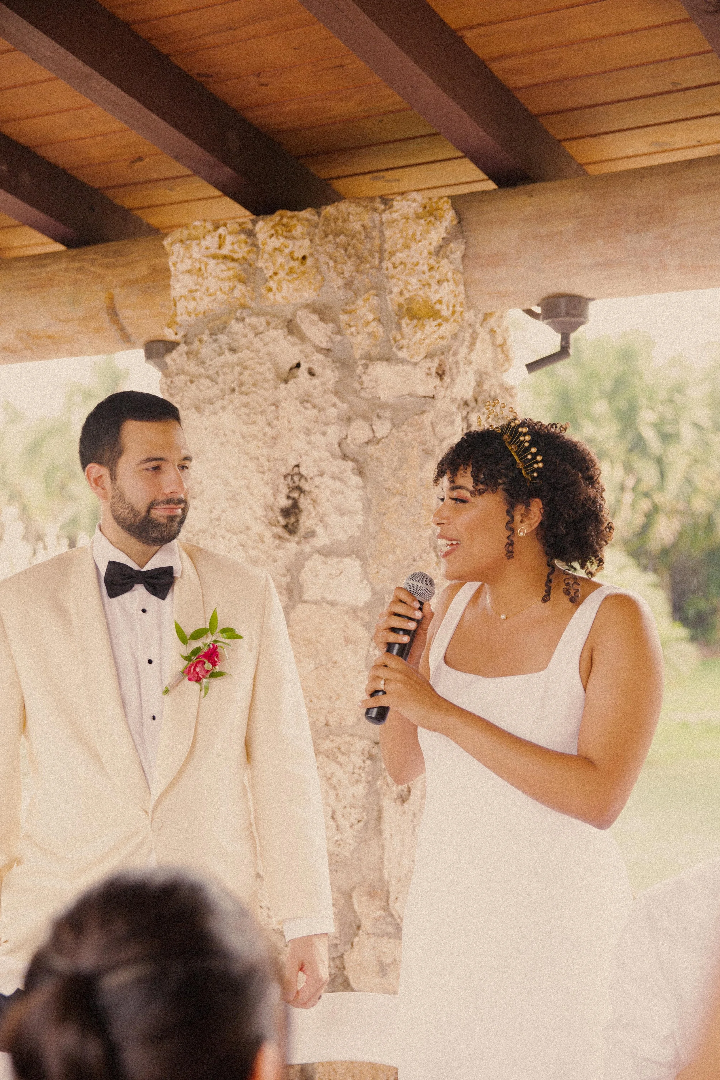 A bride and groom standing together during their wedding ceremony, with the bride speaking into a microphone, both dressed in formal attire, under a rustic stone and wood structure with greenery in the background.