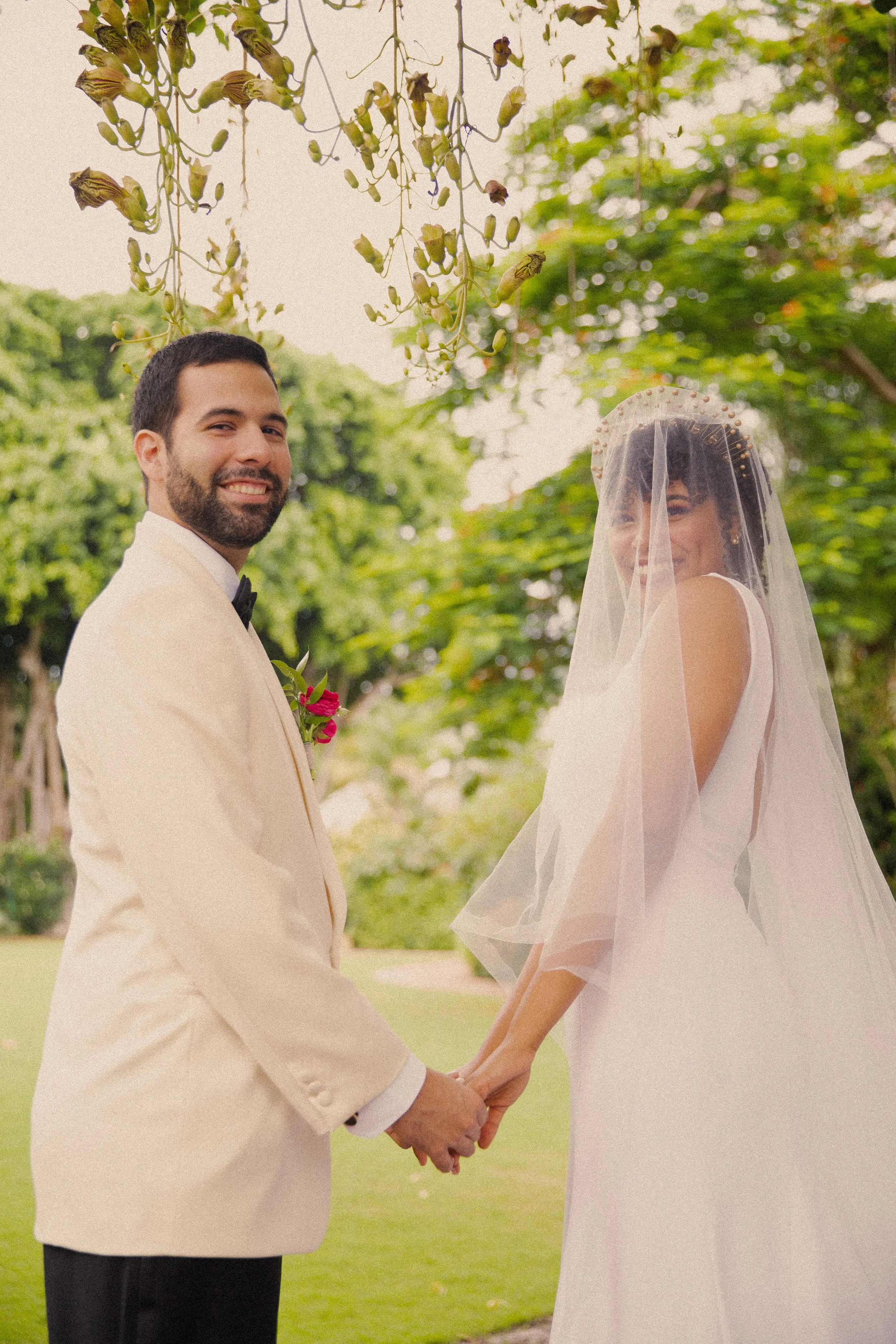 A bride and groom holding hands outdoors, smiling, with trees and greenery in the background. The bride is wearing a white wedding dress and a veil, and the groom is dressed in a light-colored suit with a dark bow tie.