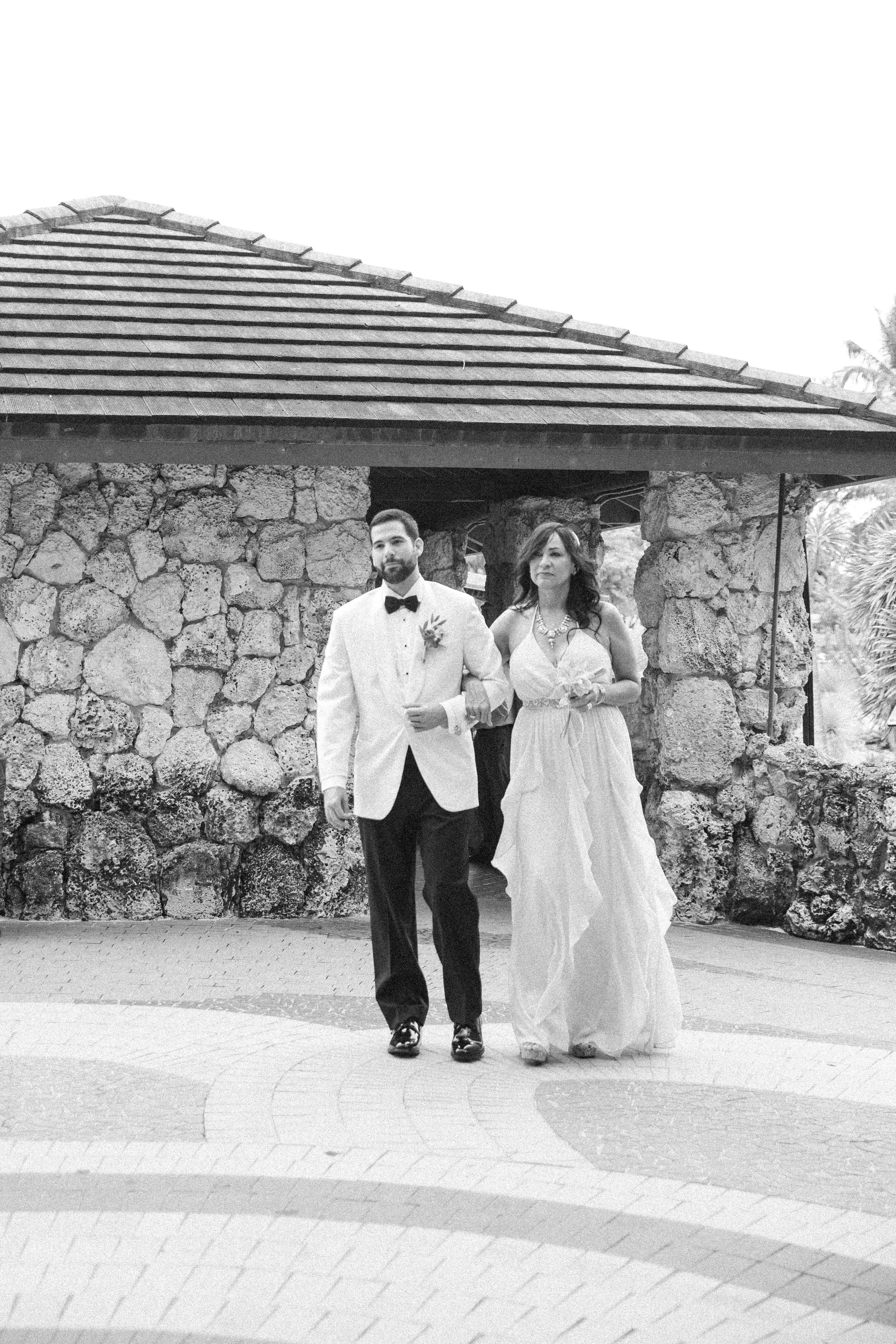 A black-and-white photograph of a bride and groom walking out of a stone building. The groom is wearing a tuxedo with a bow tie, and the bride is in a flowing white dress, holding a bouquet.