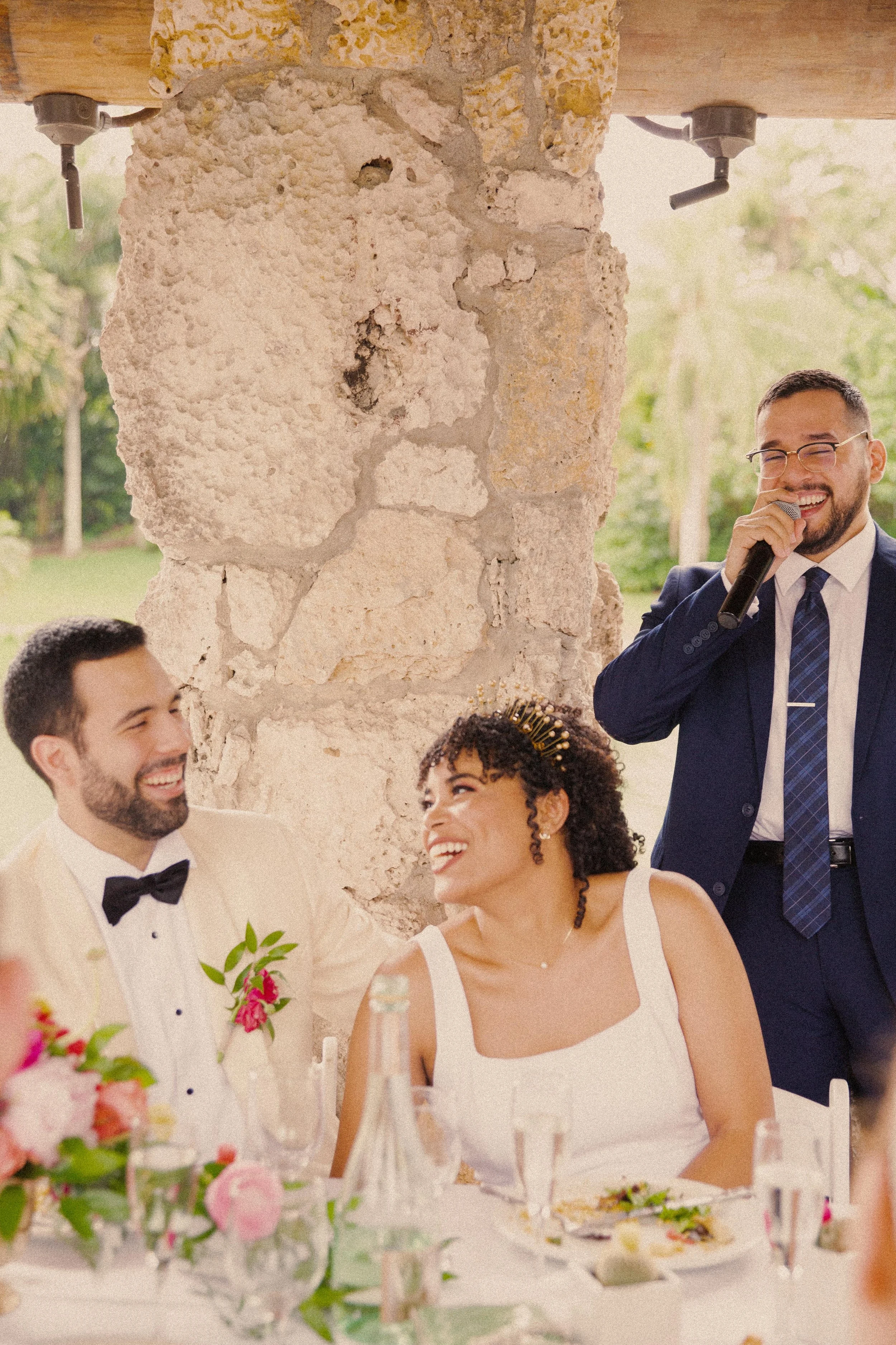 A wedding reception scene with a bride and groom sitting at a table, a man in a blue suit giving a speech with a microphone, and smiling guests, set outdoors with a stone pillar and greenery in the background.