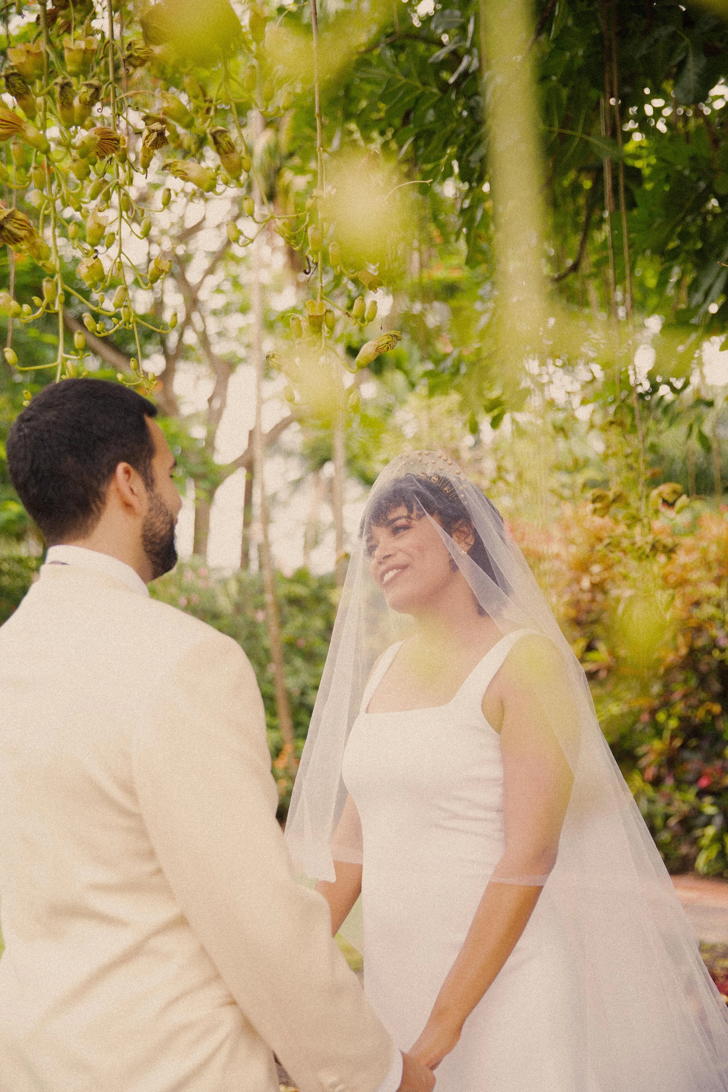 A bride and groom holding hands and gazing at each other in a garden with hanging vines and foliage.