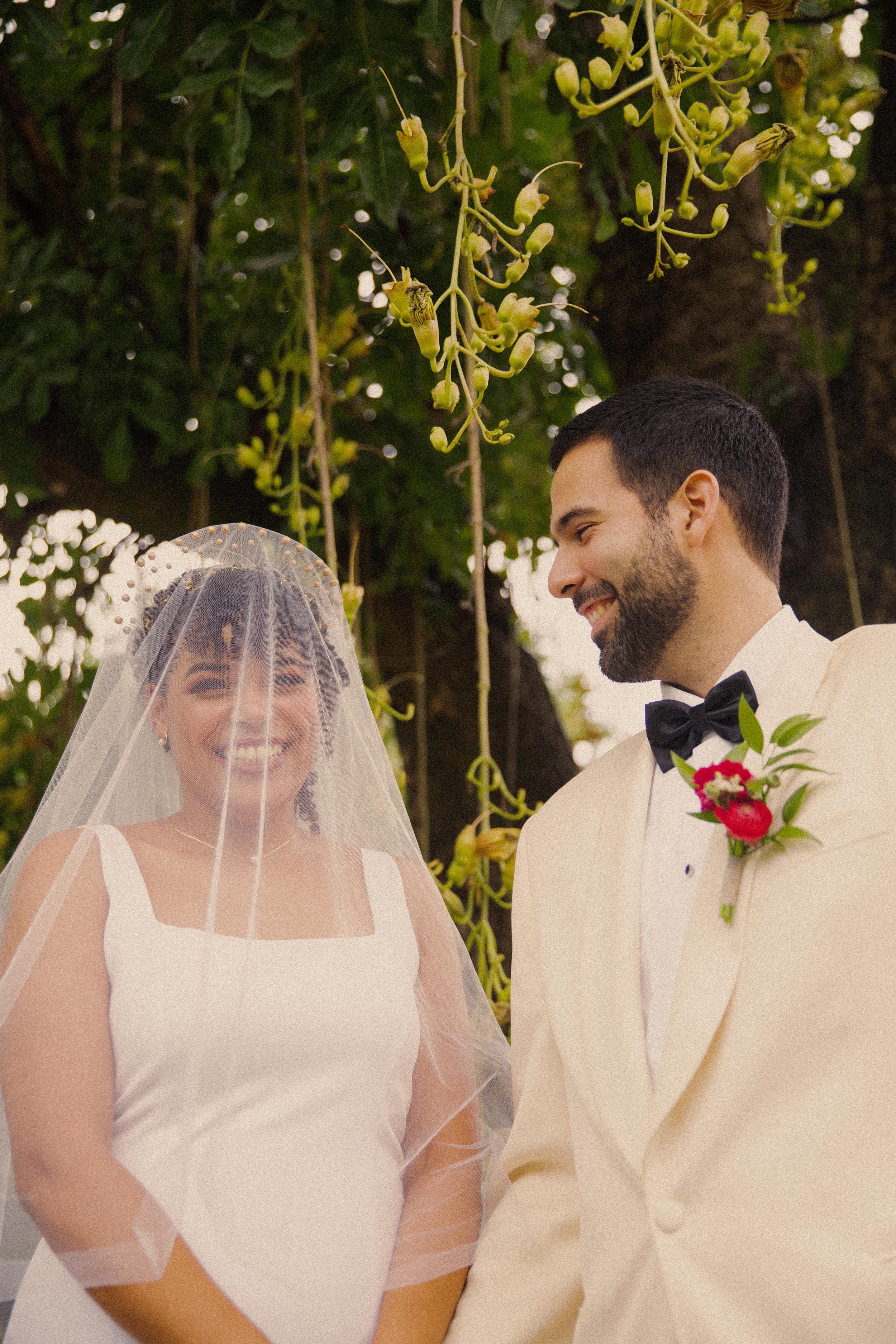 A smiling bride and groom standing outdoors under a tree with green foliage, dressed in wedding attire.