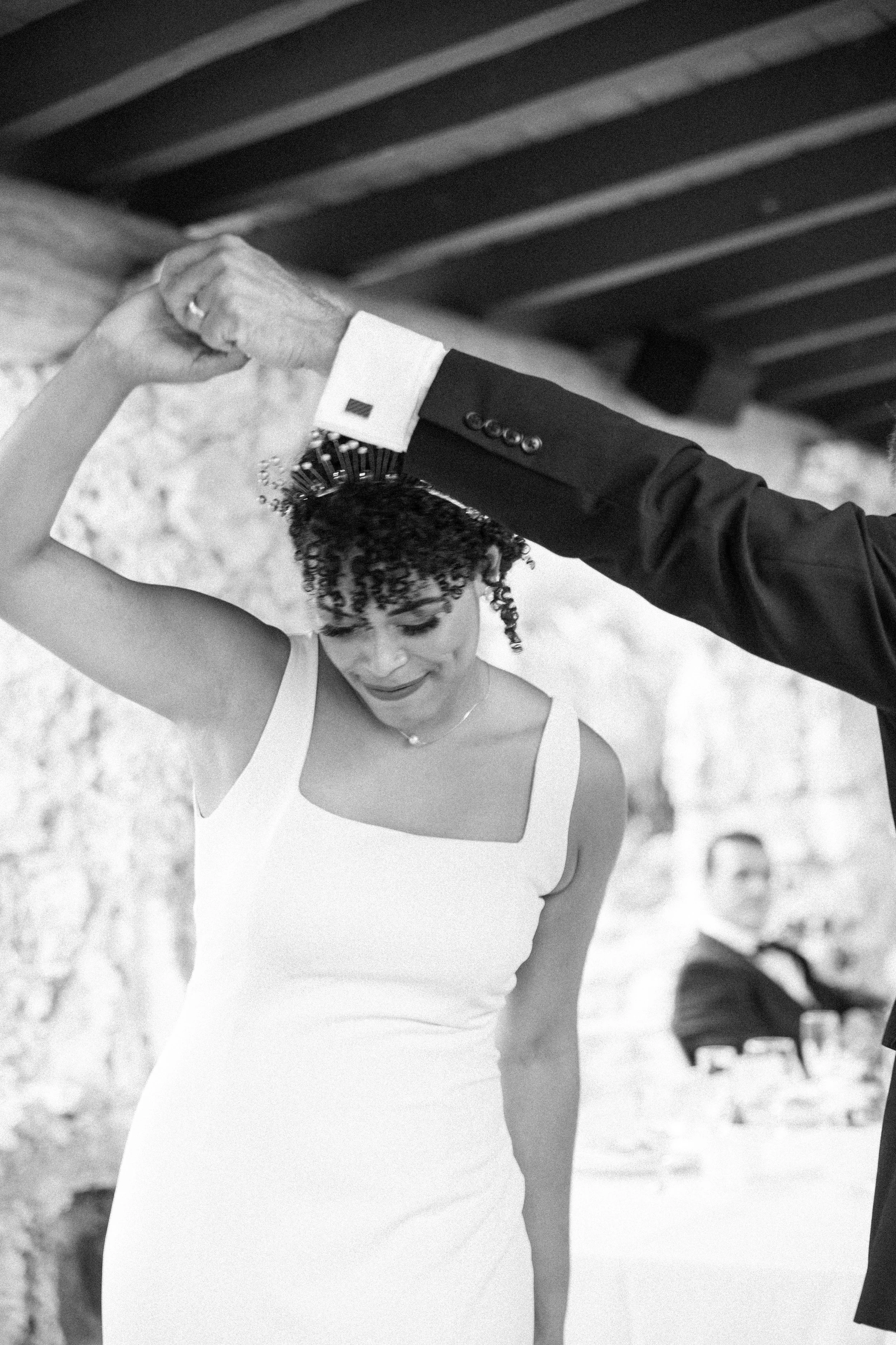 A woman in a white dress and a headpiece is dancing with a man in a dark suit at a celebration, possibly a wedding, with a guest seated in the background.