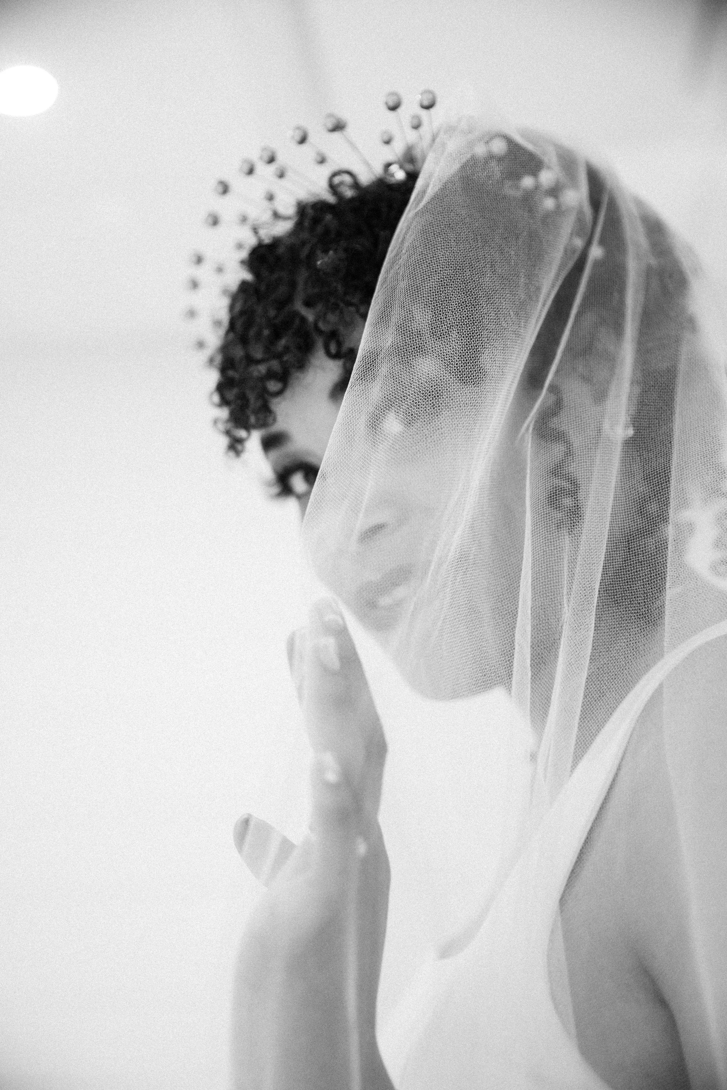 A black and white photo of a woman with short curly hair wearing a veil, looking over her shoulder, with her hand near her face.
