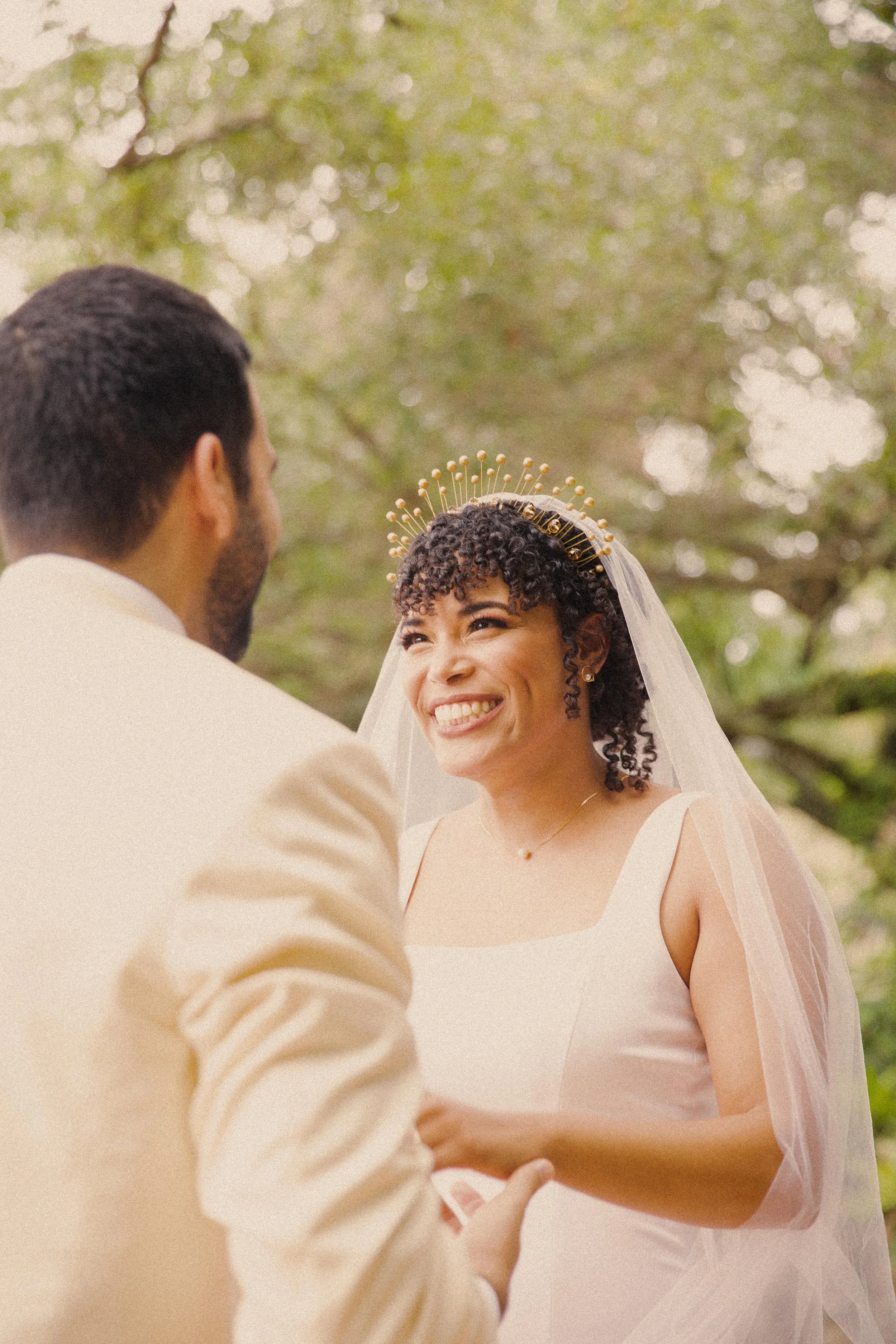 A bride and groom exchanging vows outdoors with green trees in the background. The bride is smiling and wearing a white wedding dress, a veil, and a gold headpiece, while the groom has short dark hair and a beard, dressed in a beige suit.