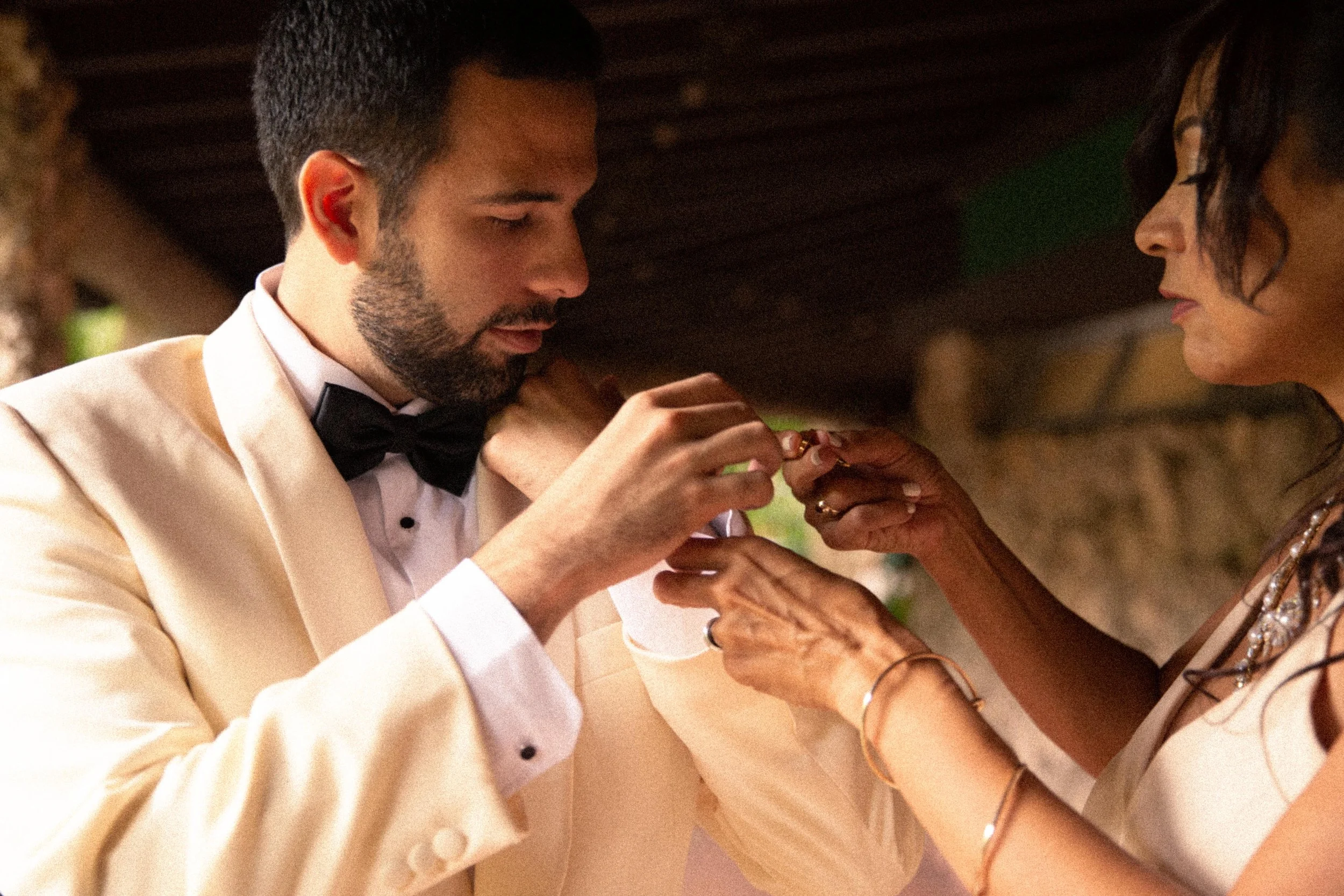 A man in a tuxedo with a black bow tie is placing a ring on a woman's finger. The woman is wearing jewelry and has dark hair. They are under a wooden structure.