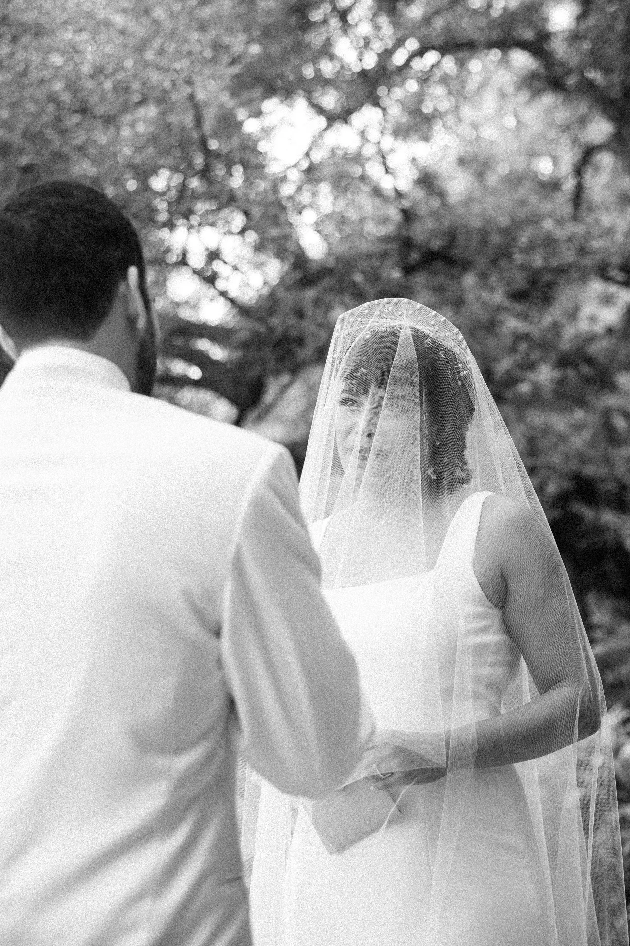 A bride wearing a wedding dress and veil looks at her groom, who is facing away from the camera, outdoors with trees and sunlight in the background.