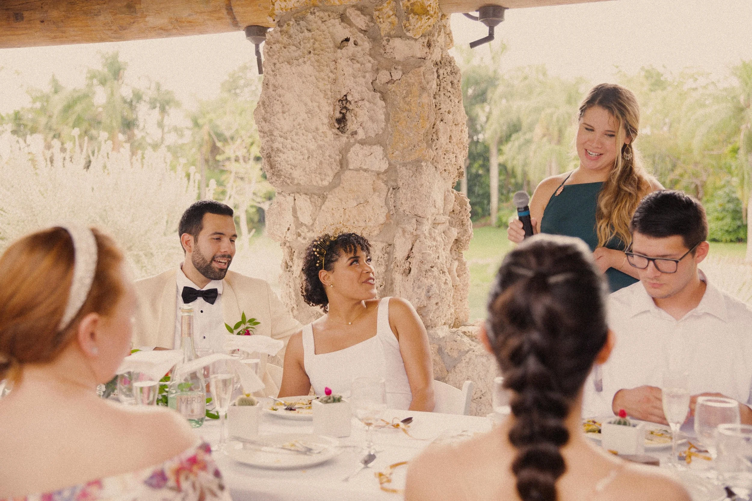 Group of people sitting at a table outdoors during a wedding reception. A woman with long red hair, wearing a floral off-shoulder dress, is visible in the foreground. In the background, a woman in a dark green dress is speaking into a microphone, whi