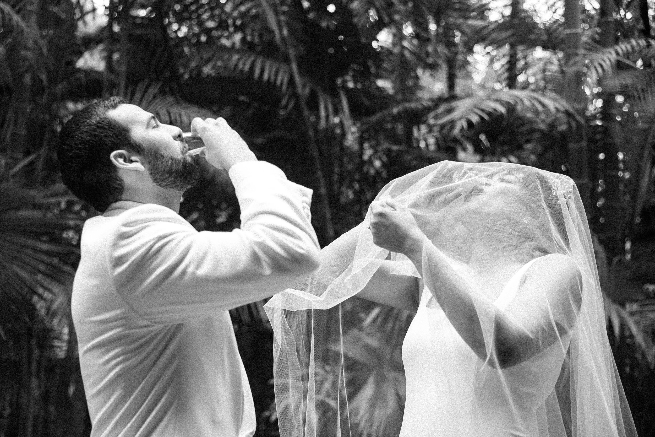 A man in a white suit drinking water and a woman in a wedding dress with a veil holding a section of her veil in a lush outdoor setting.