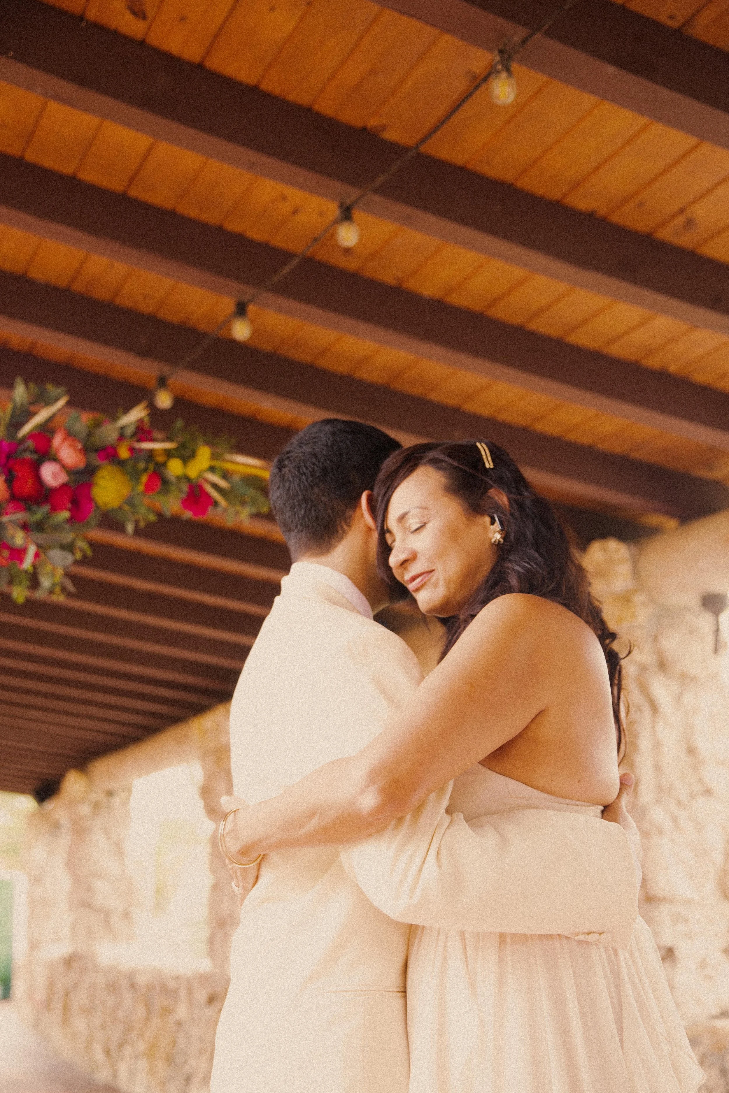 A woman and a man hugging each other, both dressed in light-colored formal attire, with flowers hanging from the ceiling and a stone wall in the background.