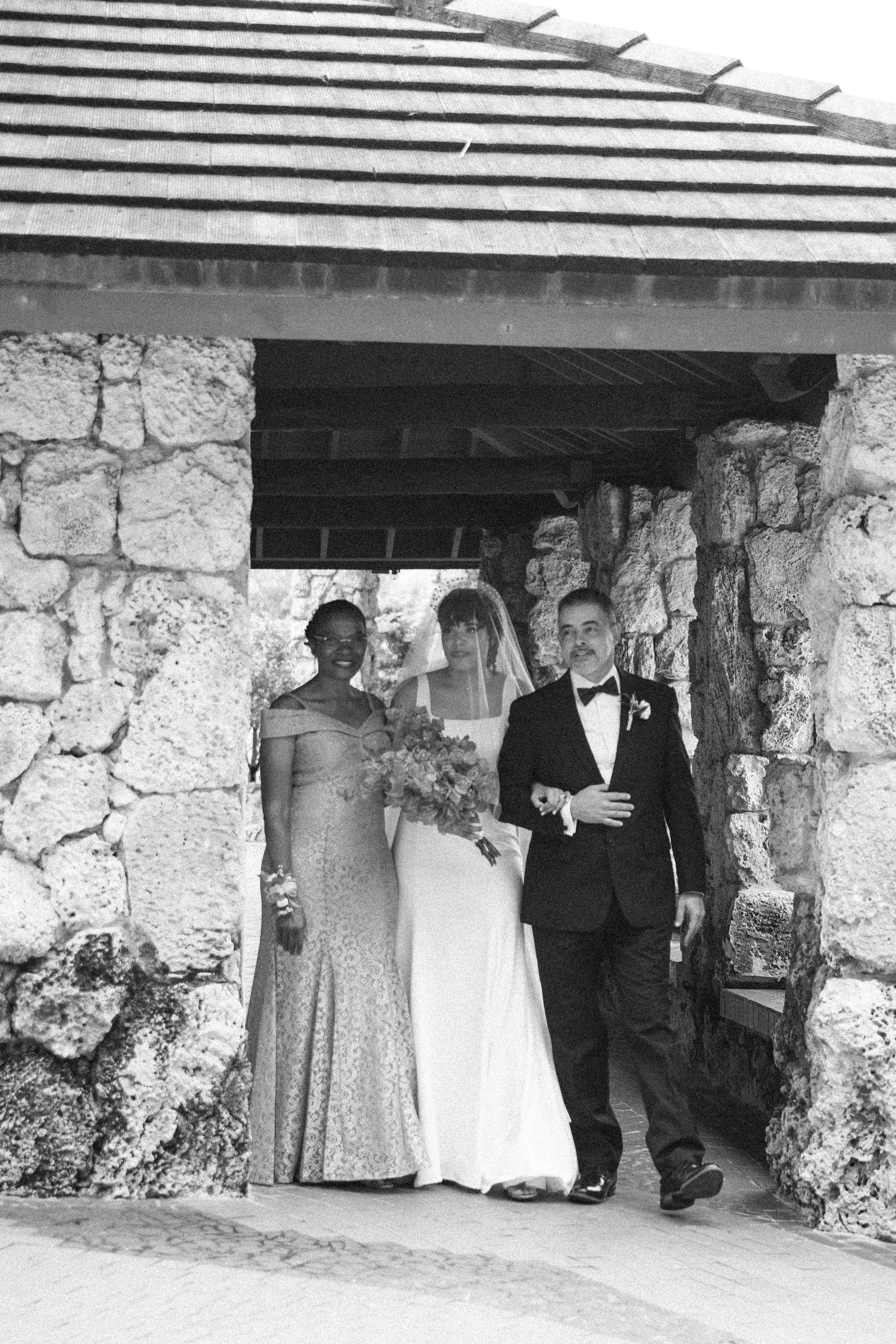 Black and white photo of a bride wearing a veil and holding a bouquet, a man in a tuxedo, and a woman in a formal gown standing together under stone archway.
