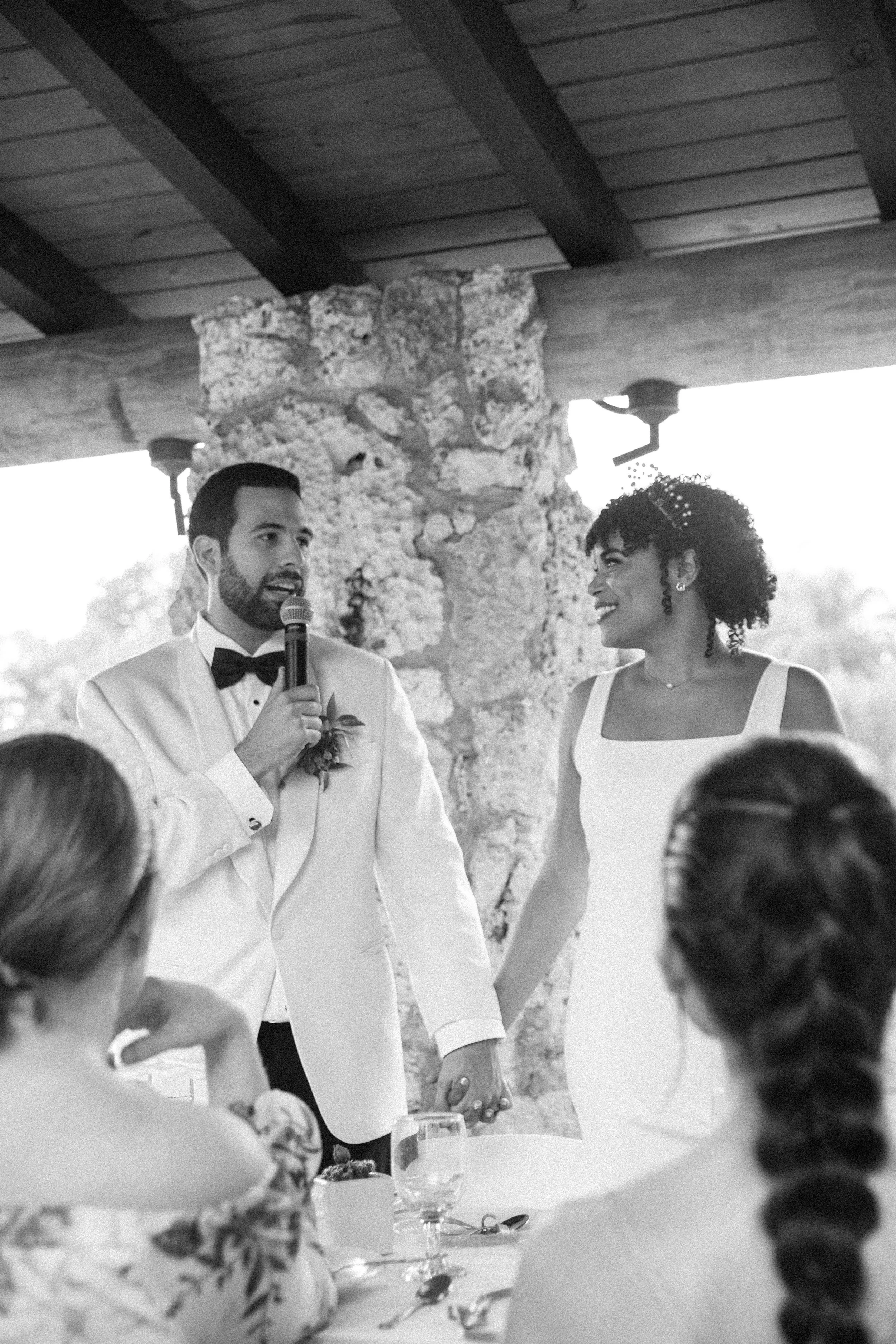 A black-and-white photo of a wedding ceremony where a groom is speaking into a microphone and holding hands with a smiling bride. The scene is inside a rustic venue with wooden ceiling beams and stone walls.