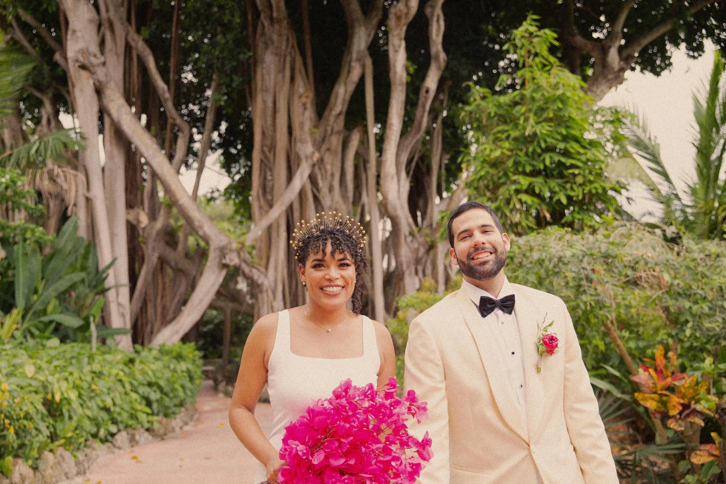 A smiling woman wearing a white dress and a gold crown holds a pink bouquet, standing next to a smiling man in a cream tuxedo with a black bow tie and a pink flower boutonniere, outdoors with large trees and lush green foliage in the background.