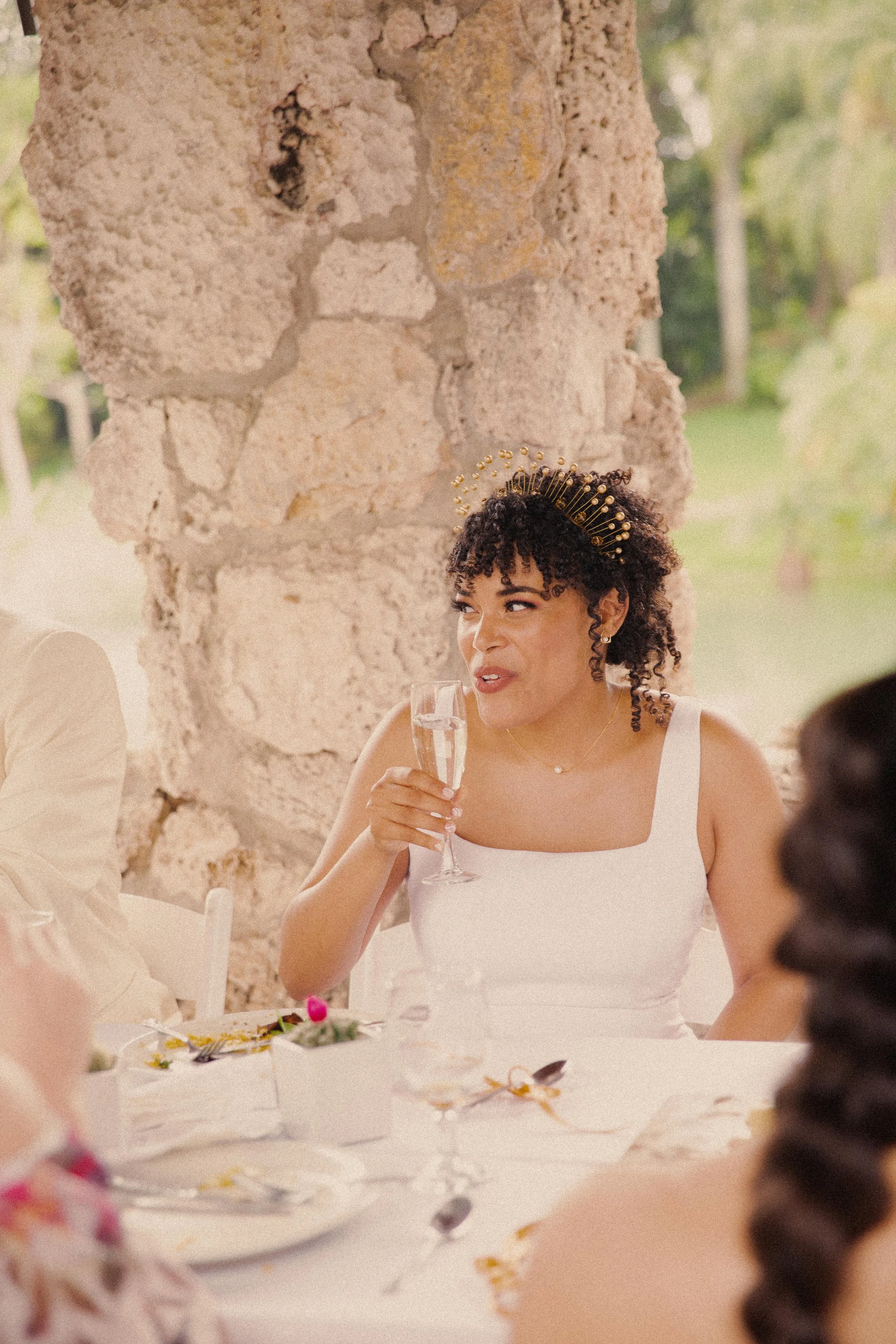 A woman with curly hair wearing a white dress and a gold headpiece, sitting at a table during a celebration, holding a glass of champagne, with a stone wall and greenery in the background.